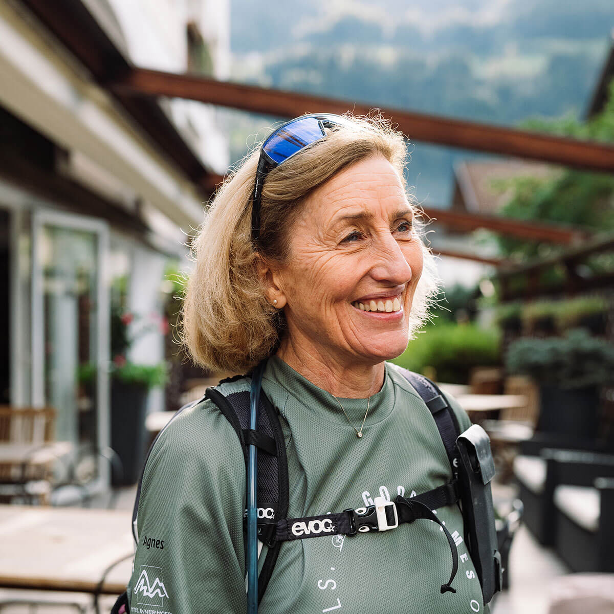 Donna anziana e sorridente con capelli corti e biondi che indossa occhiali da sole e abbigliamento da outdoor, in piedi fuori da un bar. - Hotel Innerhofer
