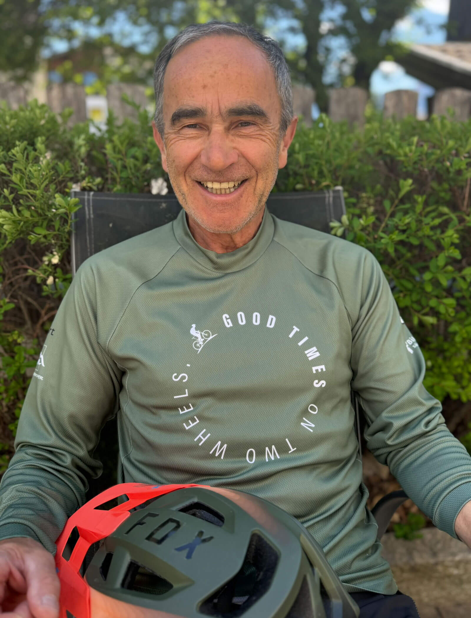 Uomo anziano sorridente con una maglietta verde Good Times on Two Wheels che tiene in mano un casco da bici arancione e nero. - Hotel Innerhofer