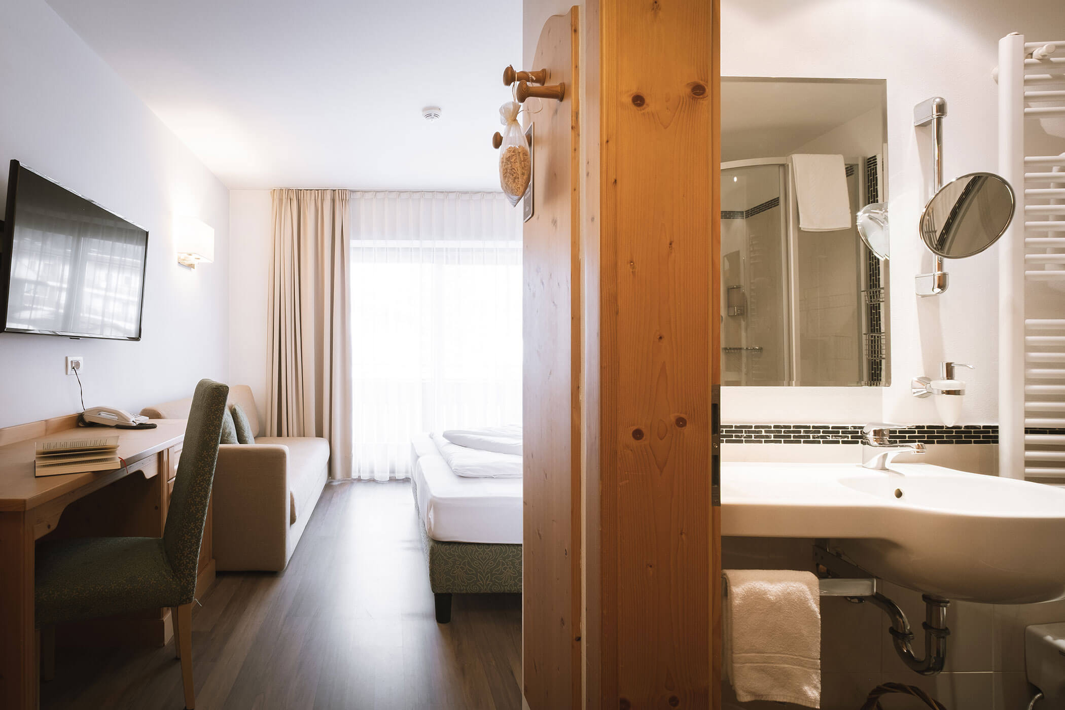 Un'accogliente camera d'albergo con letto, scrivania, divano, TV e un bagno moderno adiacente con lavandino e doccia. - Hotel Innerhofer
