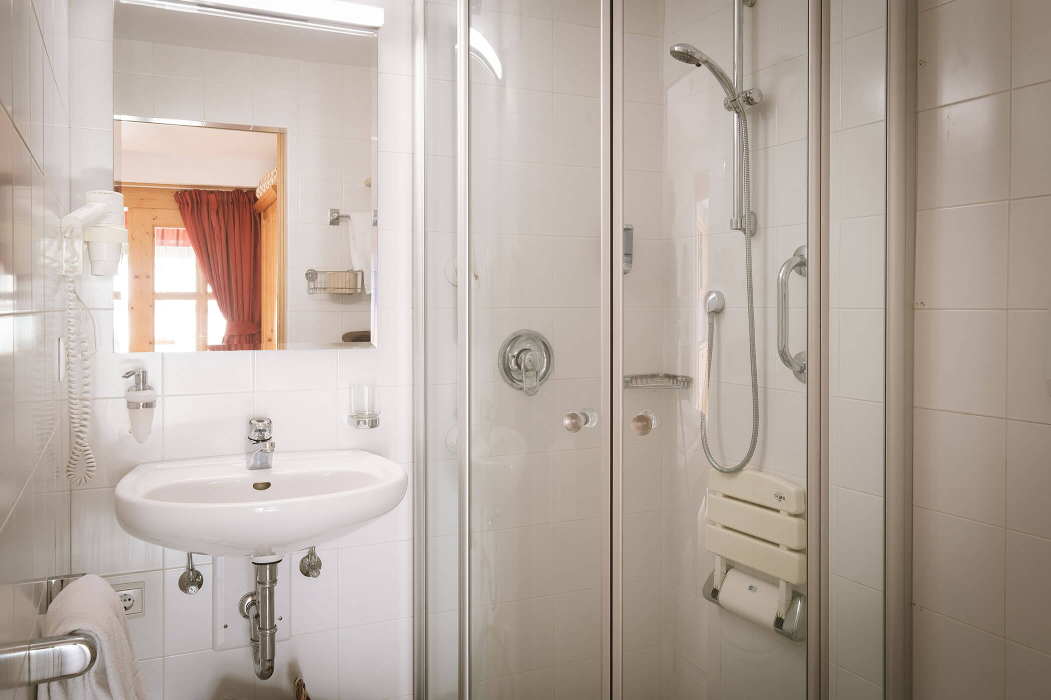 Accessible bathroom with a sink, mirror, glass shower, grab bars, and a fold-down shower seat. - Hotel Innerhofer