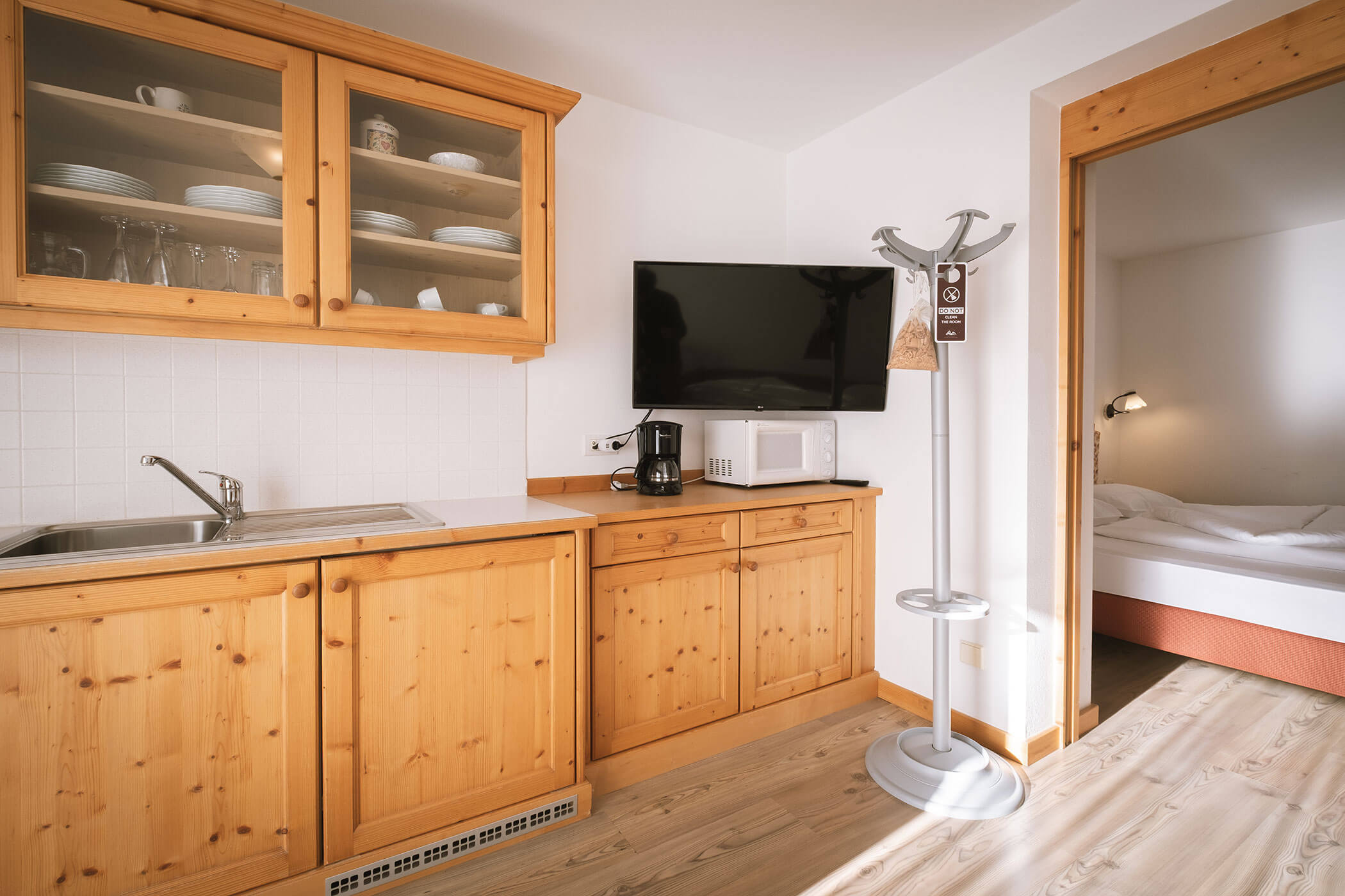Small kitchen with wooden cabinets, a sink, appliances, TV, coat rack, and a bedroom visible through door. - Hotel Innerhofer