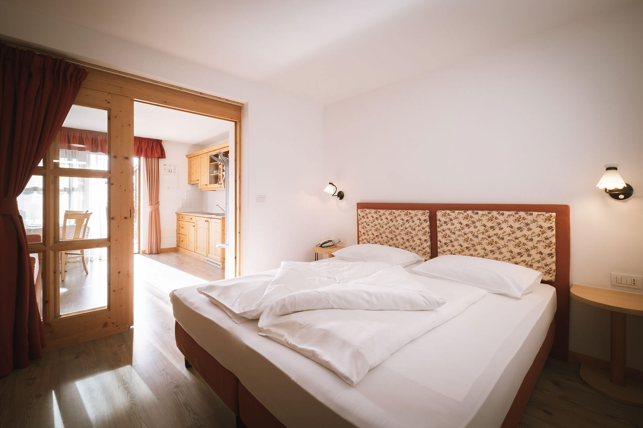 Bright hotel room with a neatly made double bed, wall lamps, and a view into a kitchen area through glass doors. - Hotel Innerhofer