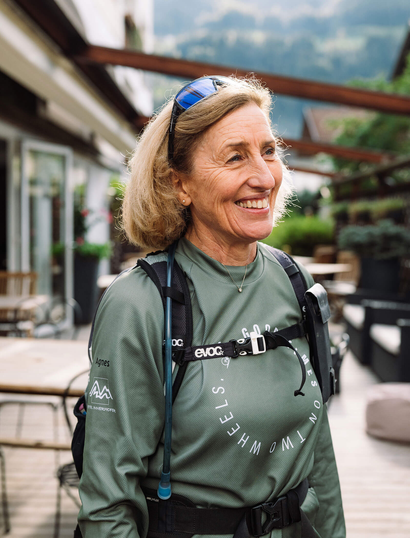 Donna sorridente in tenuta da outdoor con zaino di idratazione, in piedi su un patio con tavoli e verde. - Hotel Innerhofer