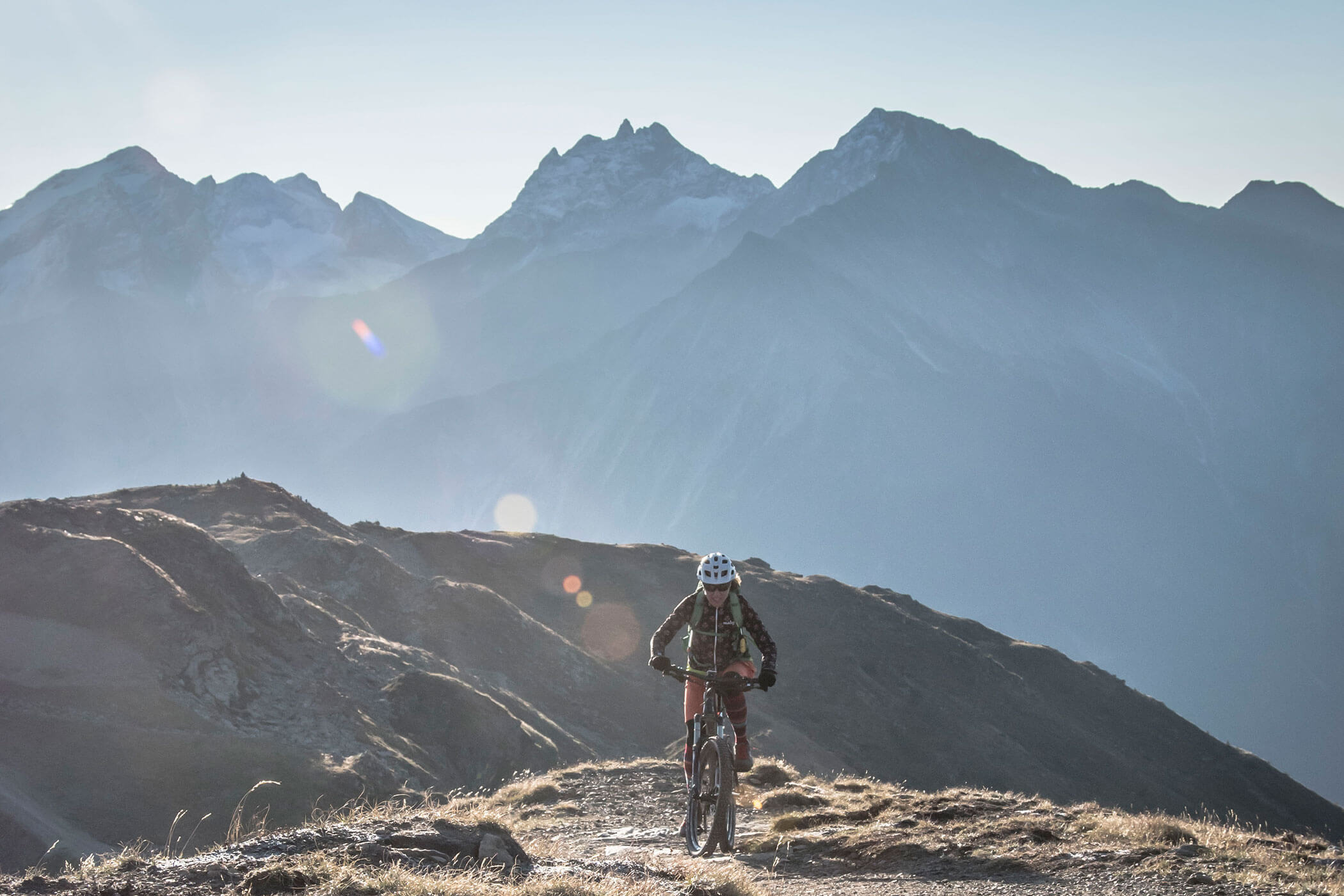 Un ciclista percorre un sentiero di montagna con alte cime aspre sullo sfondo, sotto un cielo limpido. - Hotel Innerhofer