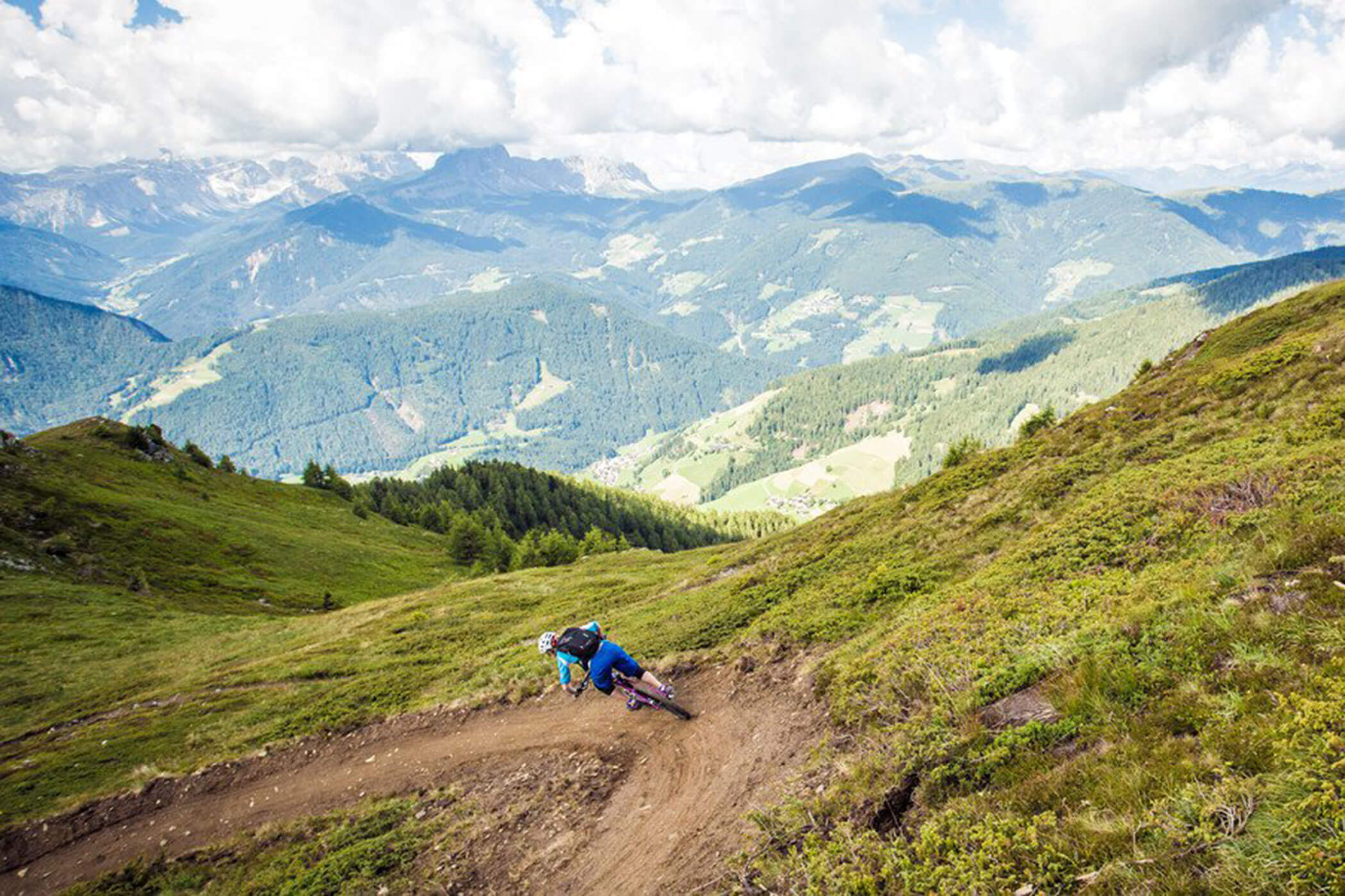 Un mountain biker percorre un tortuoso sentiero sterrato con colline e montagne verdi sullo sfondo. - Hotel Innerhofer