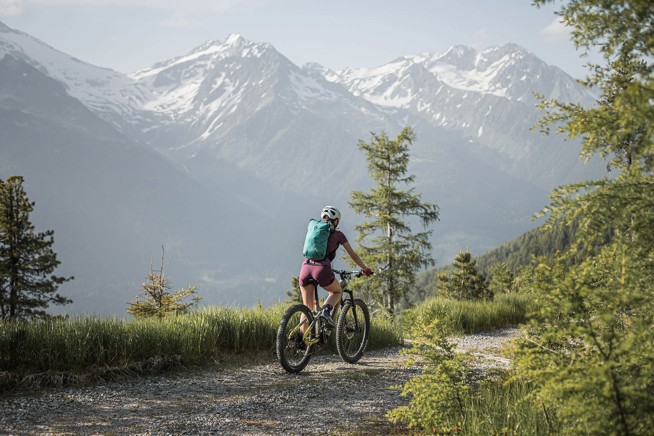 Ciclista con zaino in spalla percorre un sentiero di montagna, circondato da alberi e cime innevate sullo sfondo. - Hotel Innerhofer