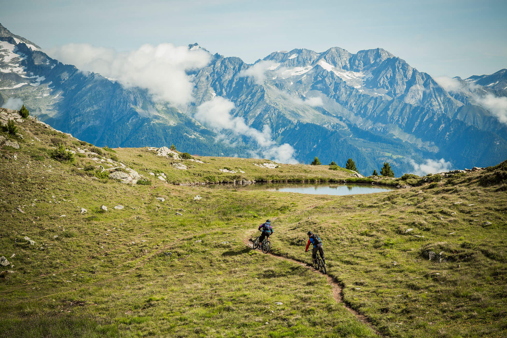 Due ciclisti percorrono un sentiero erboso di montagna con un laghetto e cime innevate sullo sfondo. - Hotel Innerhofer