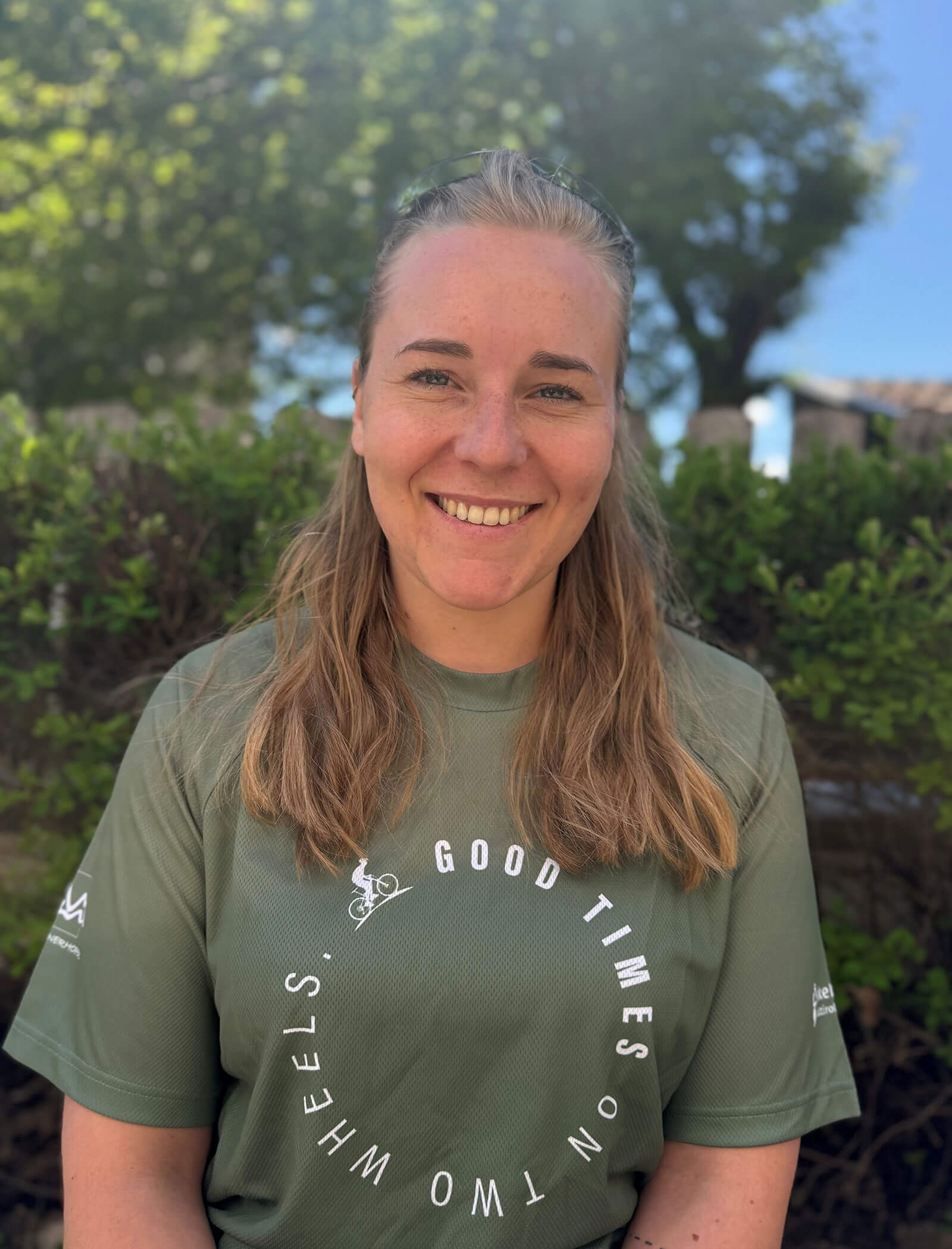 Una donna sorridente con i capelli lunghi siede all'esterno indossando una maglietta verde con la scritta Good Times On Two Wheels. - Hotel Innerhofer