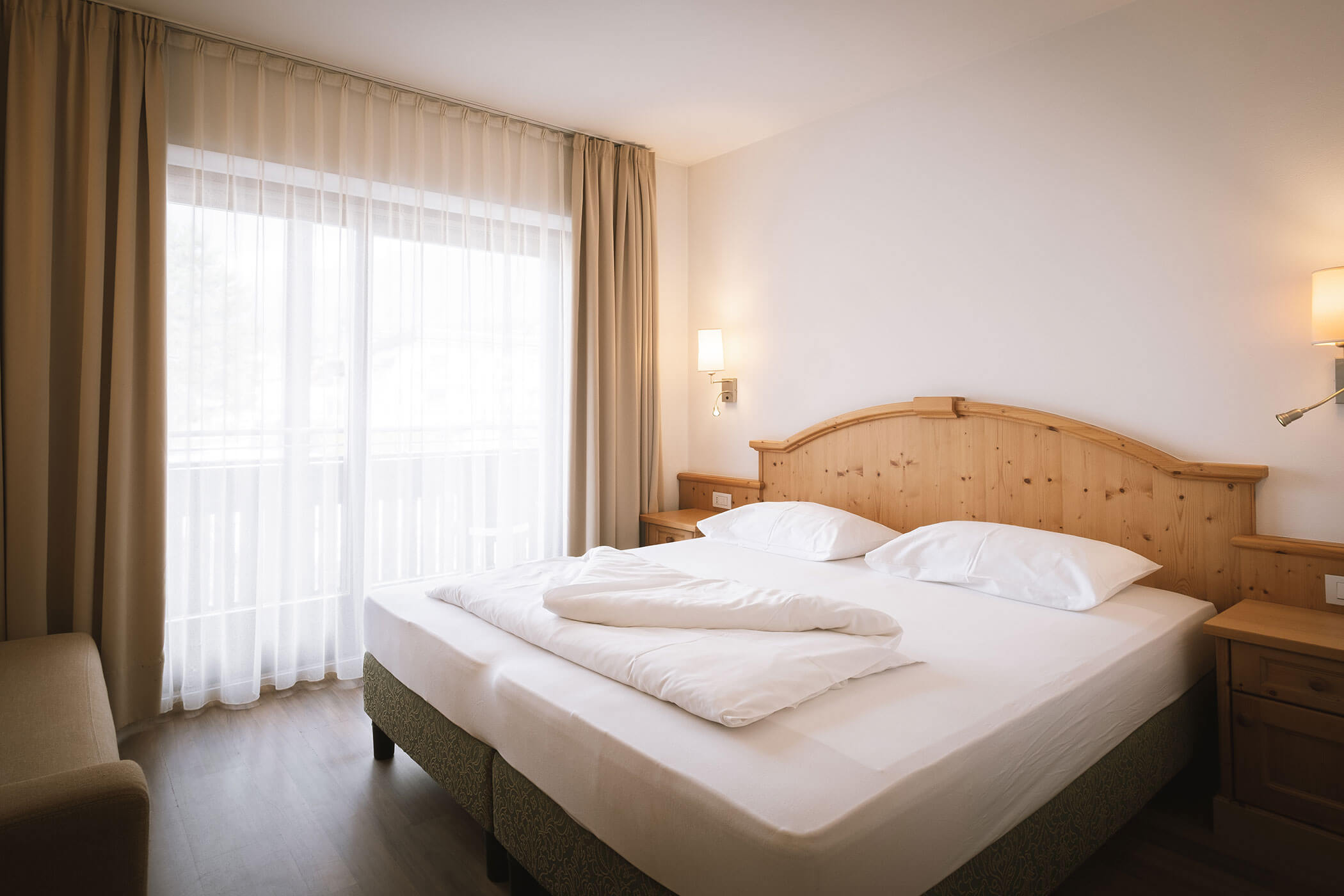 Luminosa camera d'albergo con letto matrimoniale, testiera in legno, comodini e tende trasparenti alla finestra. - Hotel Innerhofer