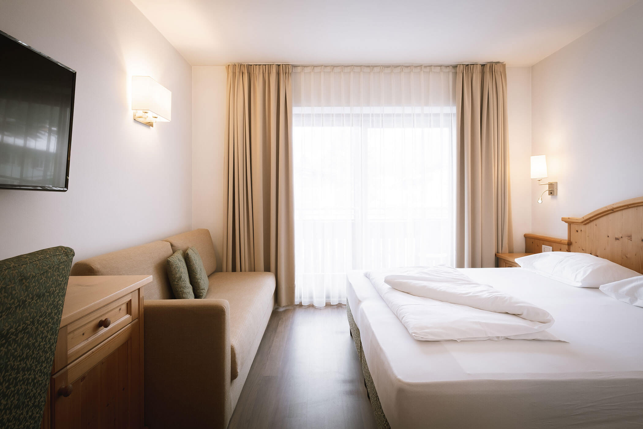 Luminosa camera d'albergo con letto matrimoniale, divano, TV, mobili in legno e ampia finestra con tende trasparenti. - Hotel Innerhofer