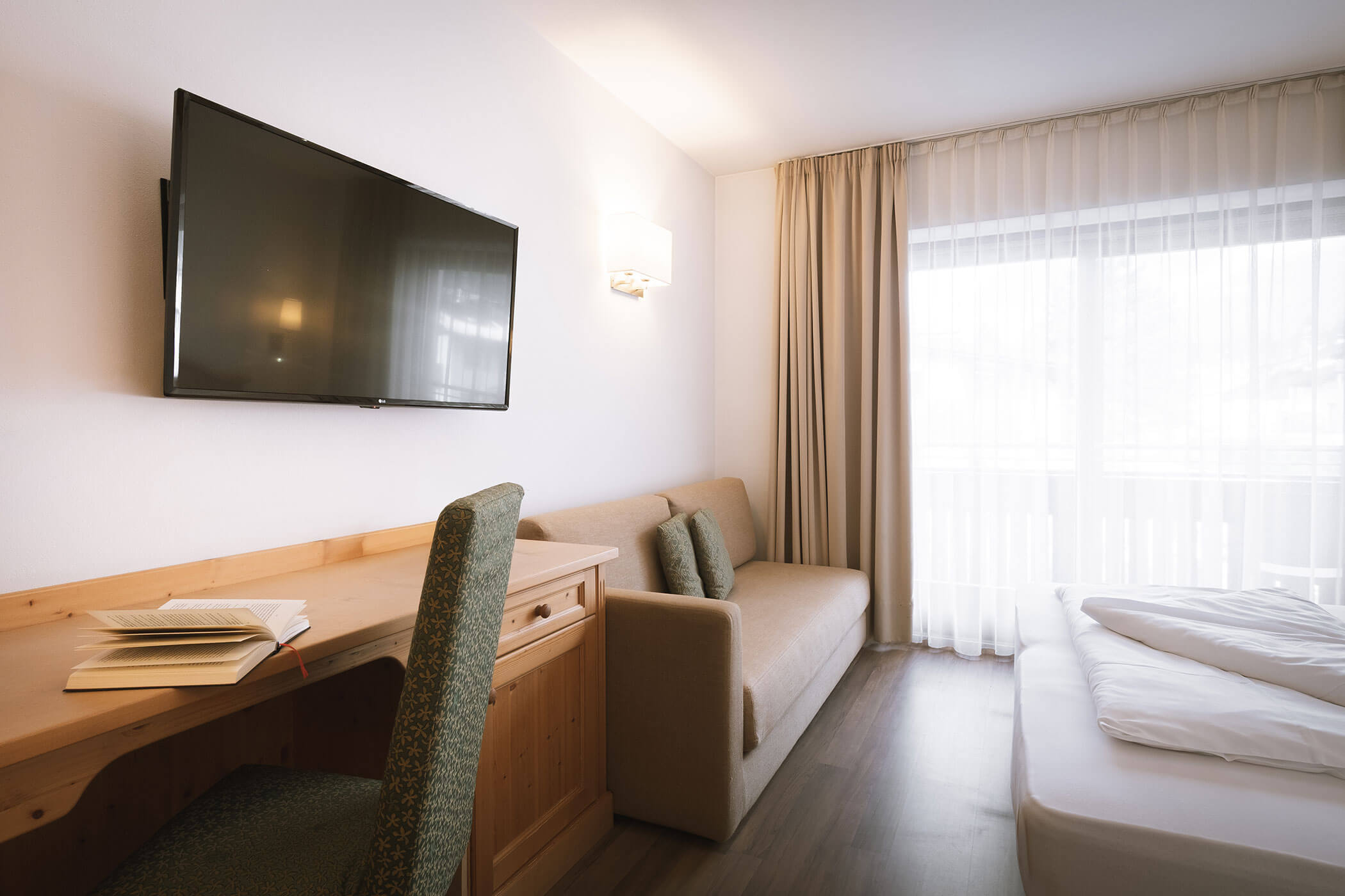 Camera d'albergo con letto, divano, scrivania, sedia, TV a parete e ampia finestra con tende trasparenti. - Hotel Innerhofer