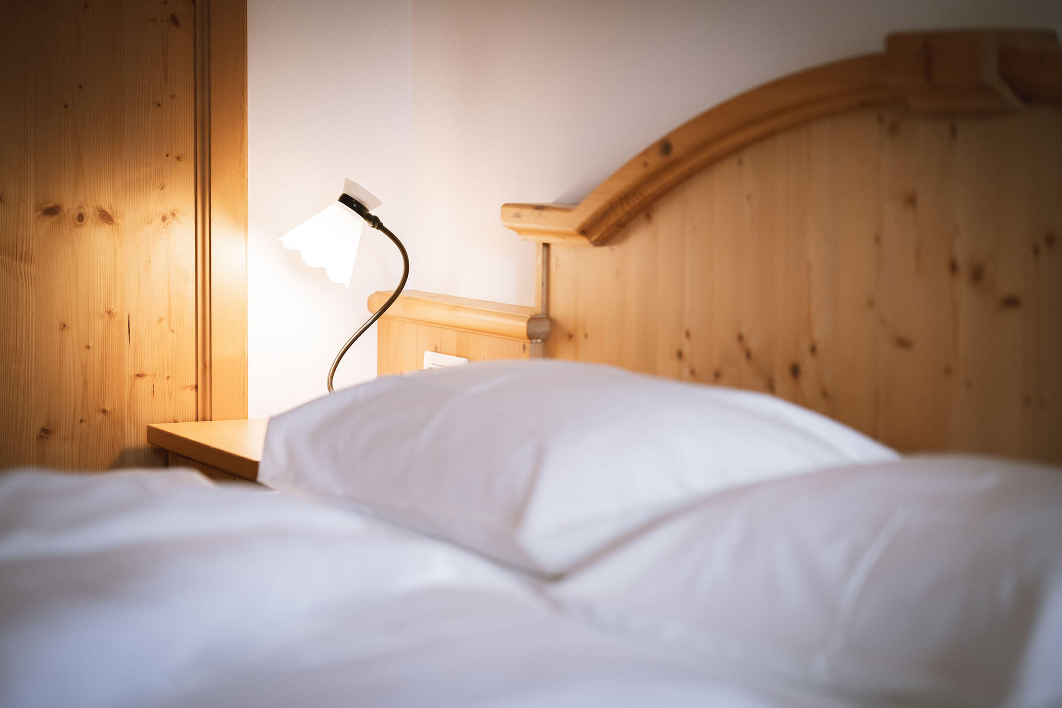 Primo piano di un letto ben fatto con cuscini bianchi e testiera in legno, accanto a una lampada da comodino accesa. - Hotel Innerhofer