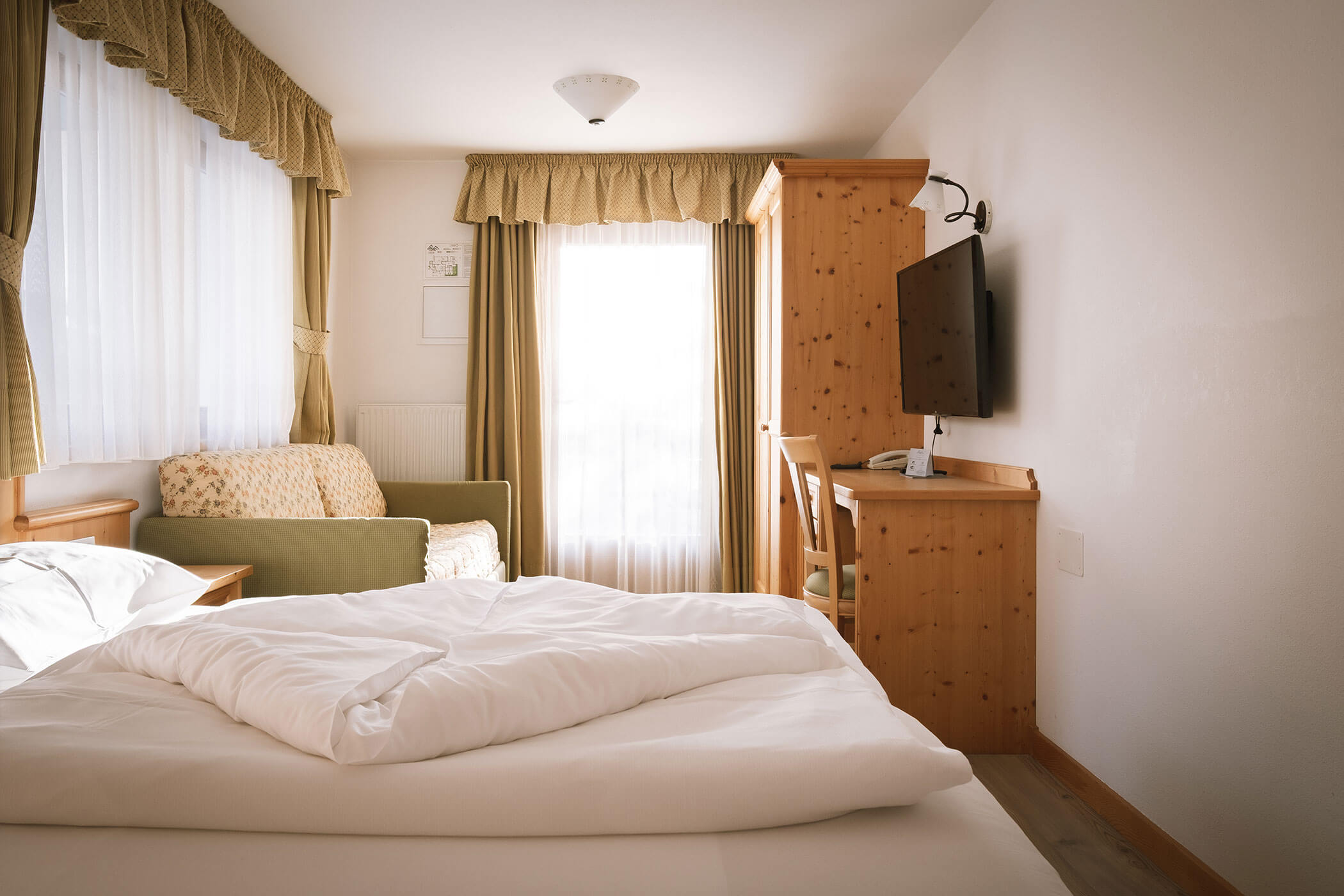 Accogliente camera d'albergo con letto matrimoniale, mobili in legno, divano, scrivania, TV e luce del sole attraverso le finestre. - Hotel Innerhofer