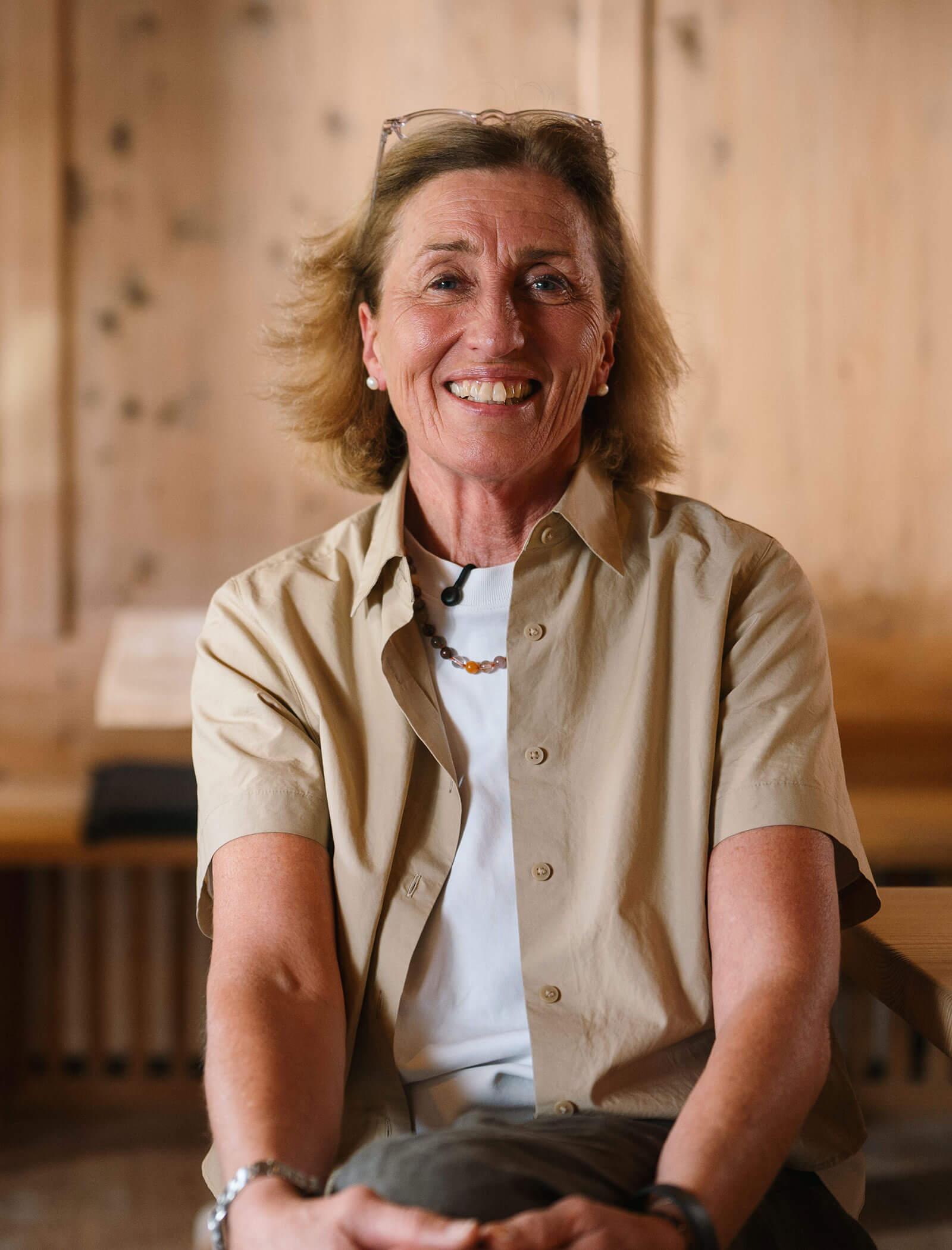 Smiling woman with short hair wearing a beige shirt and white top, sitting indoors against a wooden background. - Hotel Innerhofer