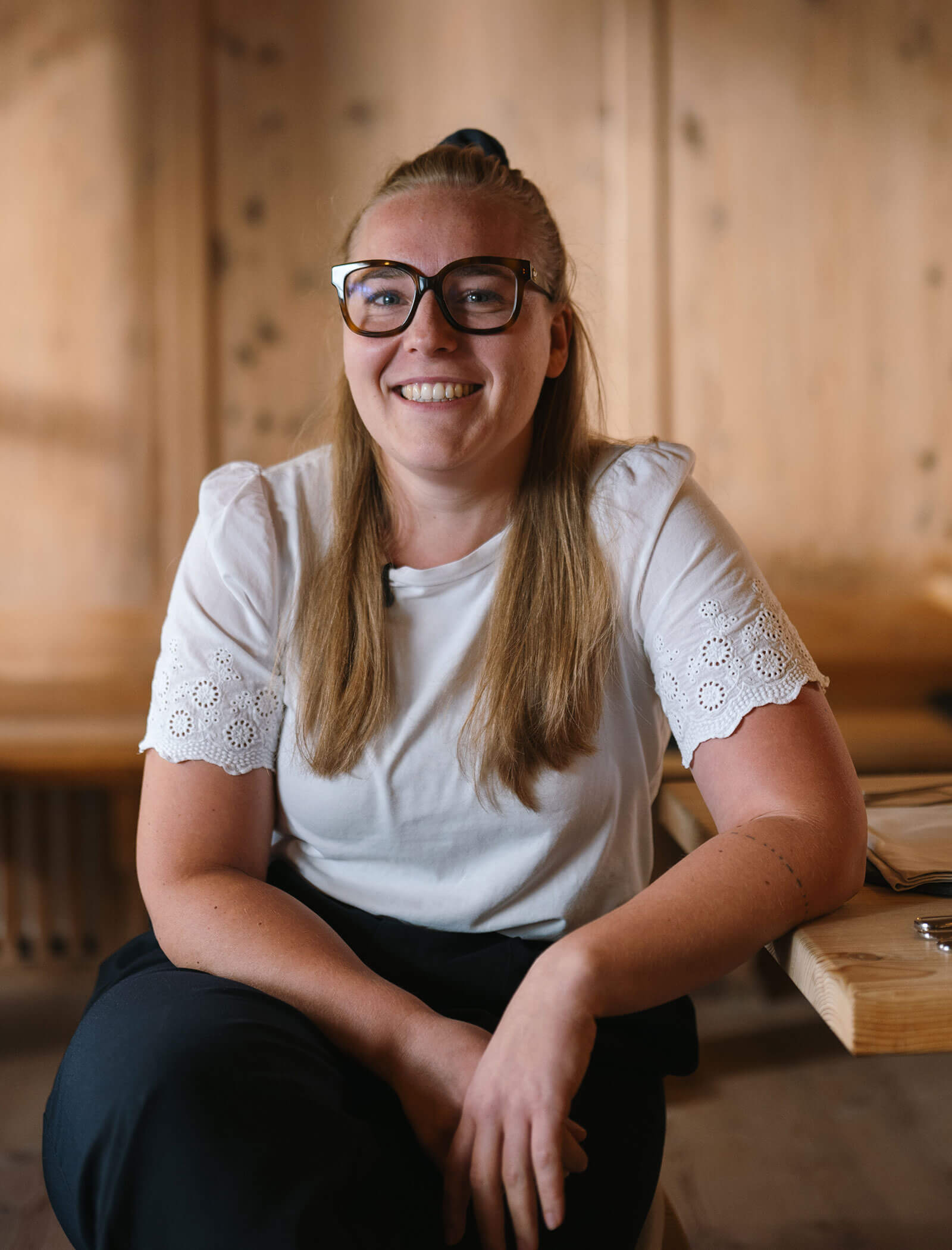 Smiling woman with glasses and long blonde hair sitting at a wooden table in a warmly lit room. - Hotel Innerhofer