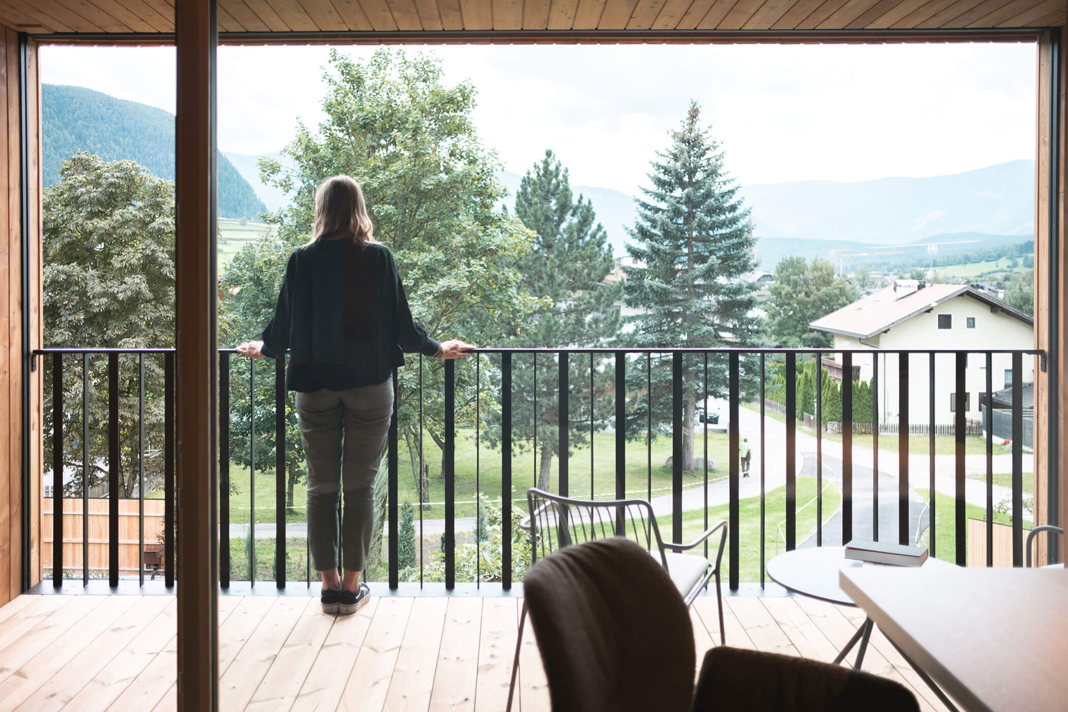 Person standing on a balcony, looking at a scenic view of trees, houses, and distant mountains. - Hotel Innerhofer