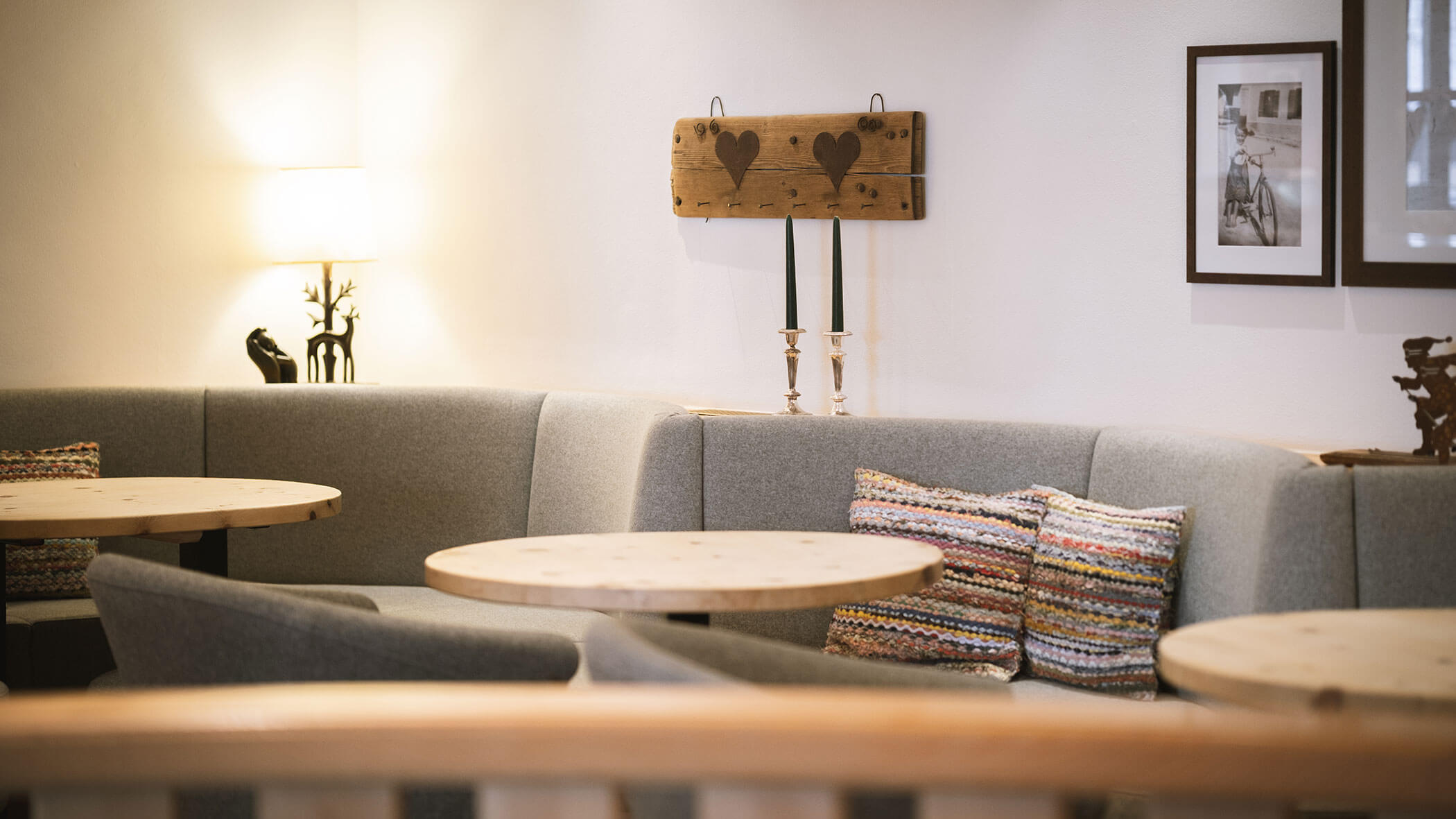 Cozy cafe corner with grey sofas, round wooden tables, and decorative pillows, softly lit by a lamp. - Hotel Innerhofer