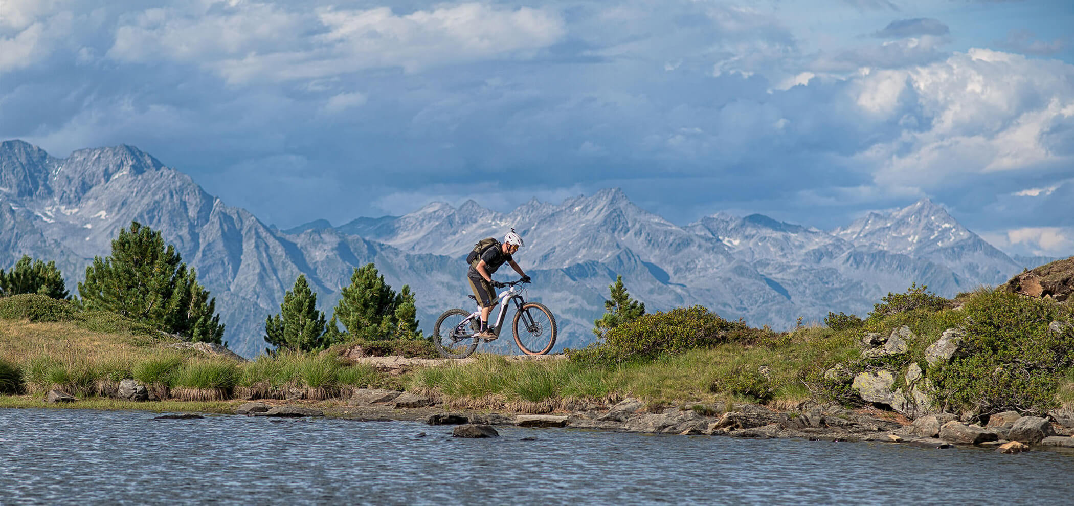 Un ciclista pedala in mountain bike su un sentiero vicino a un lago, con alte montagne e alberi sullo sfondo. - Hotel Innerhofer