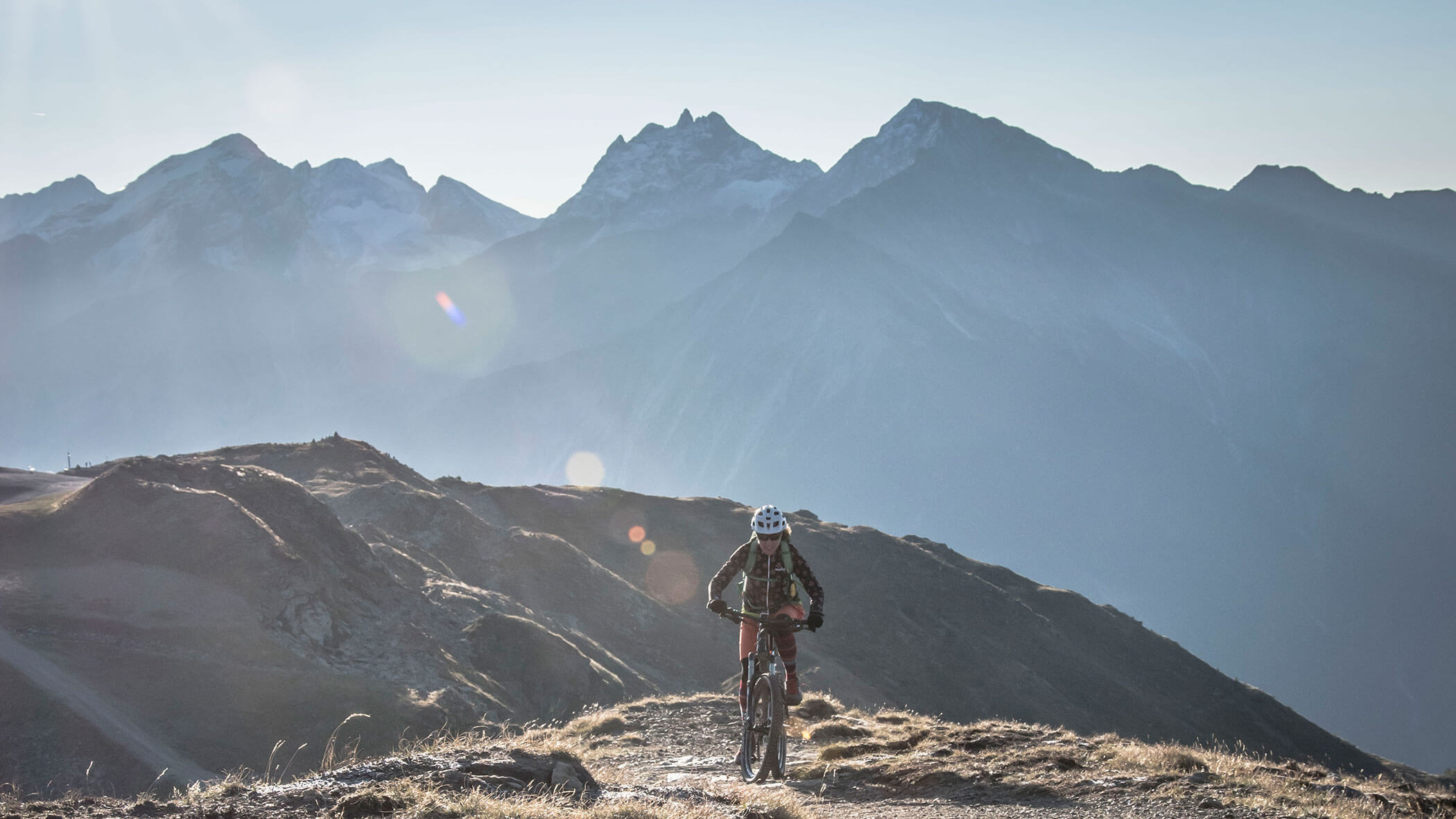 A mountain biker rides along a ridge trail with tall, sunlit mountains in the background. - Hotel Innerhofer