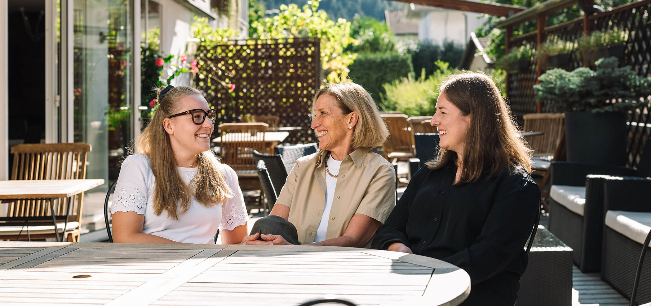 Three women sit together at an outdoor café table, smiling and chatting in the sunlight. - Hotel Innerhofer
