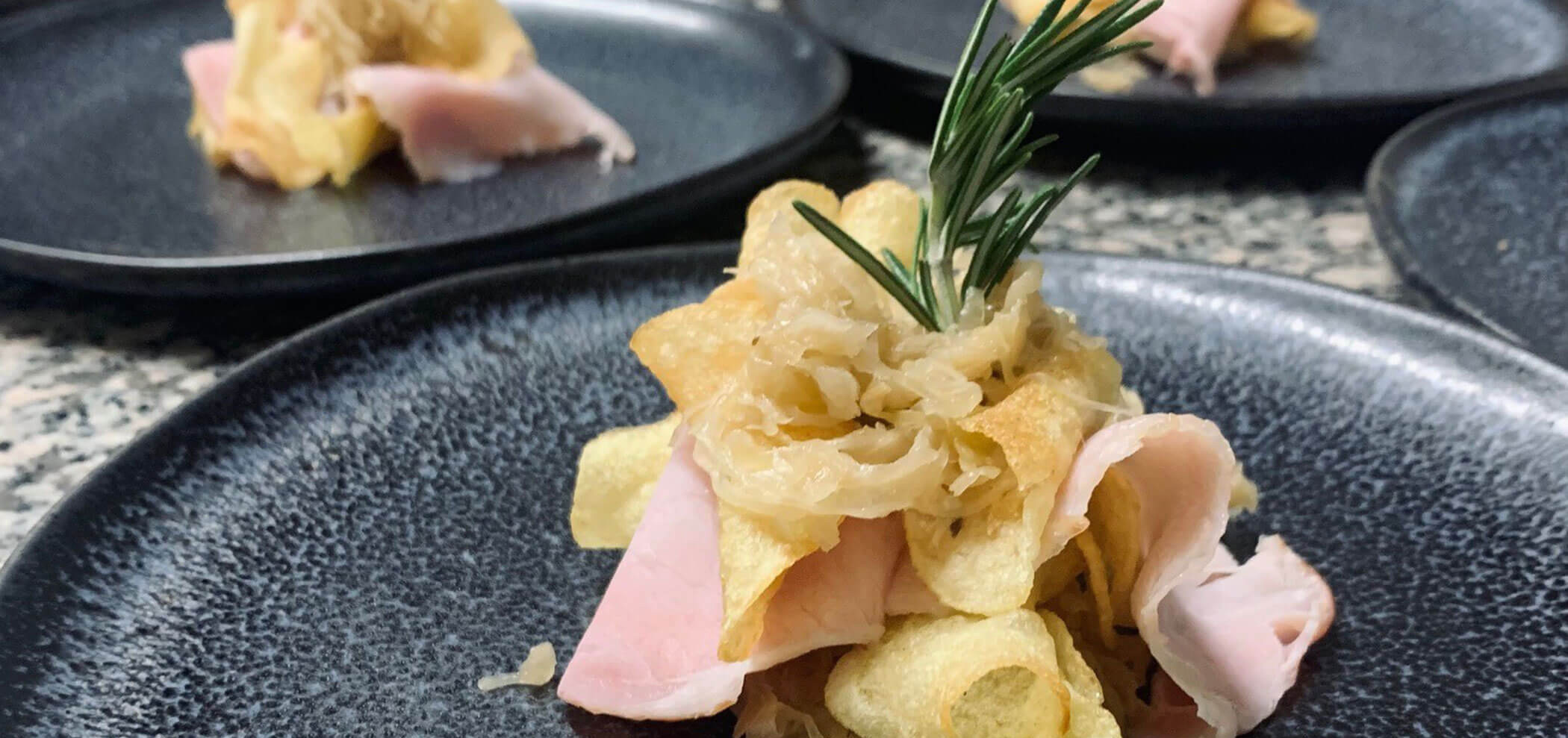 Ein schwarzer Teller mit Schinken, Kartoffelchips, Sauerkraut und einem Zweig Rosmarin, der als Vorspeise serviert wird. - Hotel Innerhofer