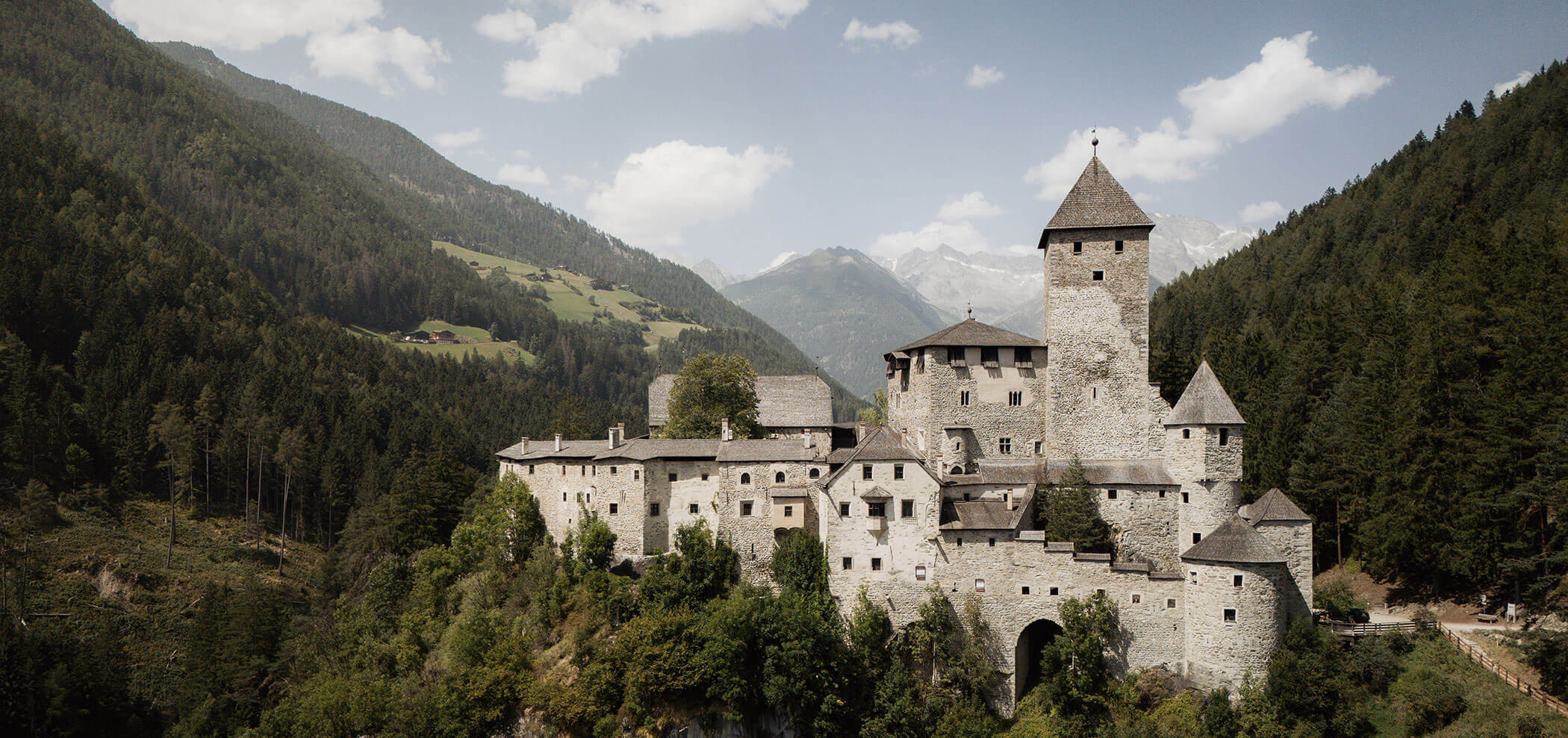 Medieval stone castle surrounded by forested mountains under a partly cloudy sky. - Hotel Innerhofer