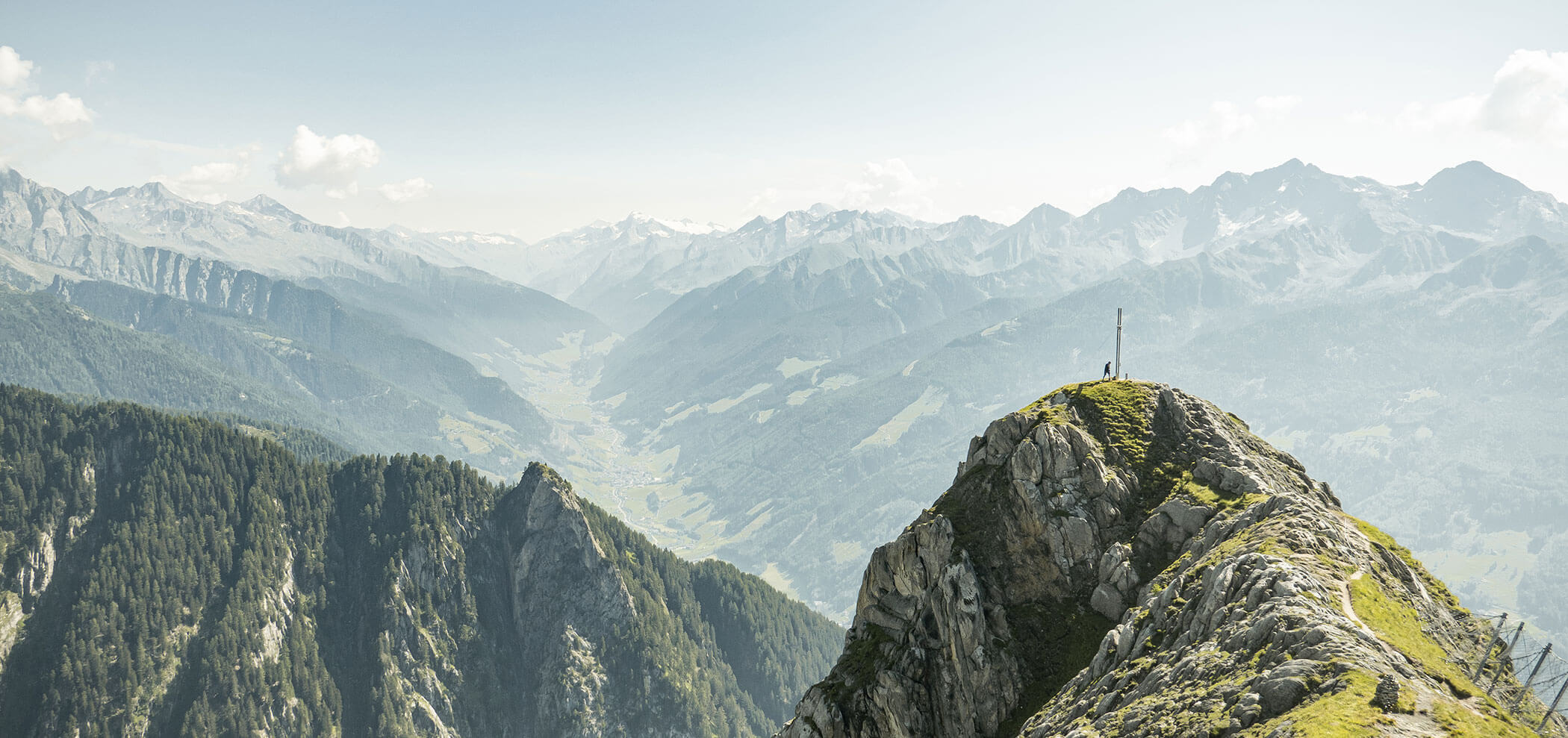 Un escursionista si trova sul picco di una montagna rocciosa che domina una valle verdeggiante e le lontane montagne innevate. - Hotel Innerhofer