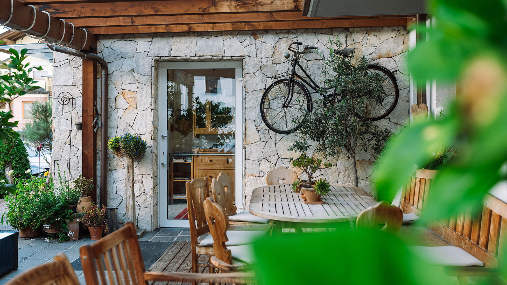 A cozy patio with wooden furniture, potted plants, and a bicycle mounted on a stone wall. - Hotel Innerhofer