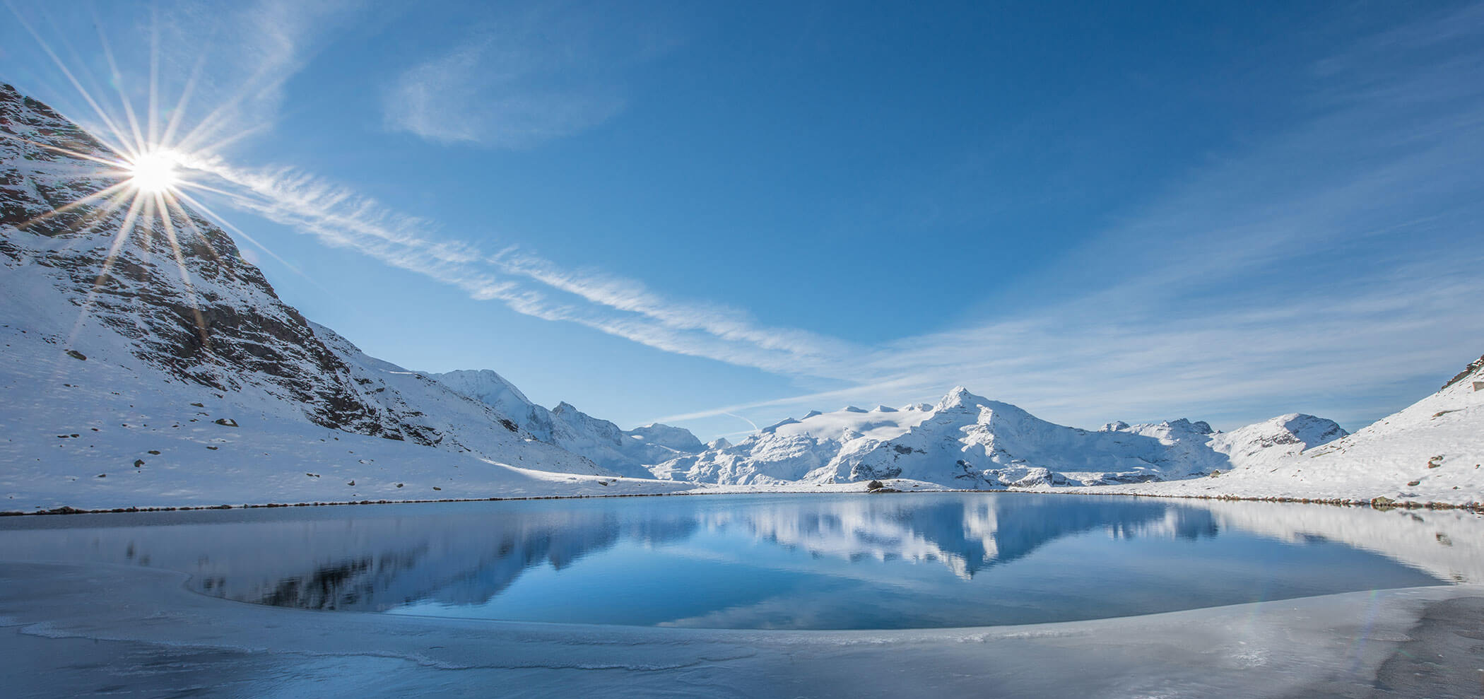 Snowy mountains reflected in a calm, partially frozen lake with the sun shining over a peak and blue sky above. - Hotel Innerhofer