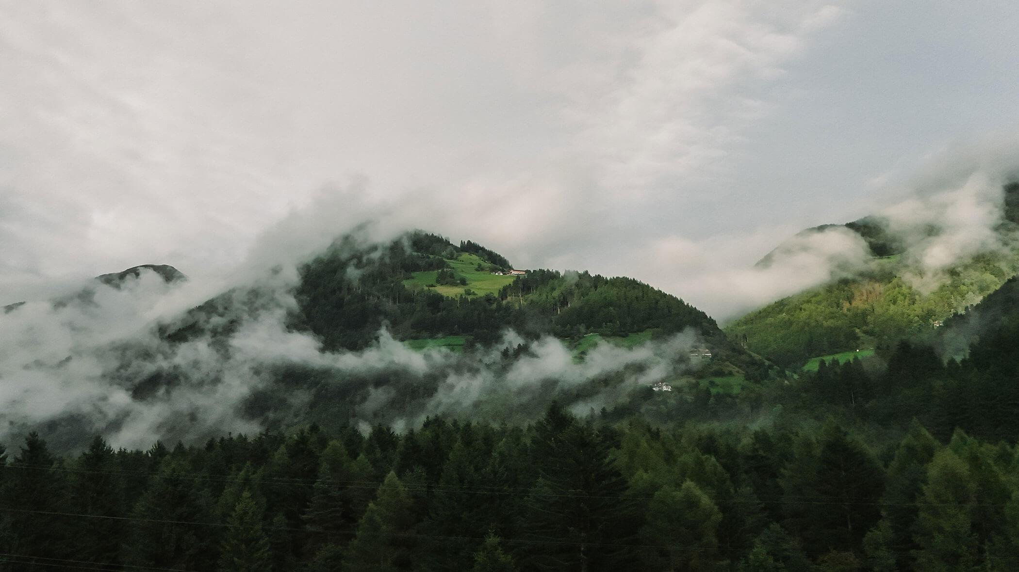 Misty clouds drift over green forested mountains with patches of sunlight and grassy hills. - Hotel Innerhofer