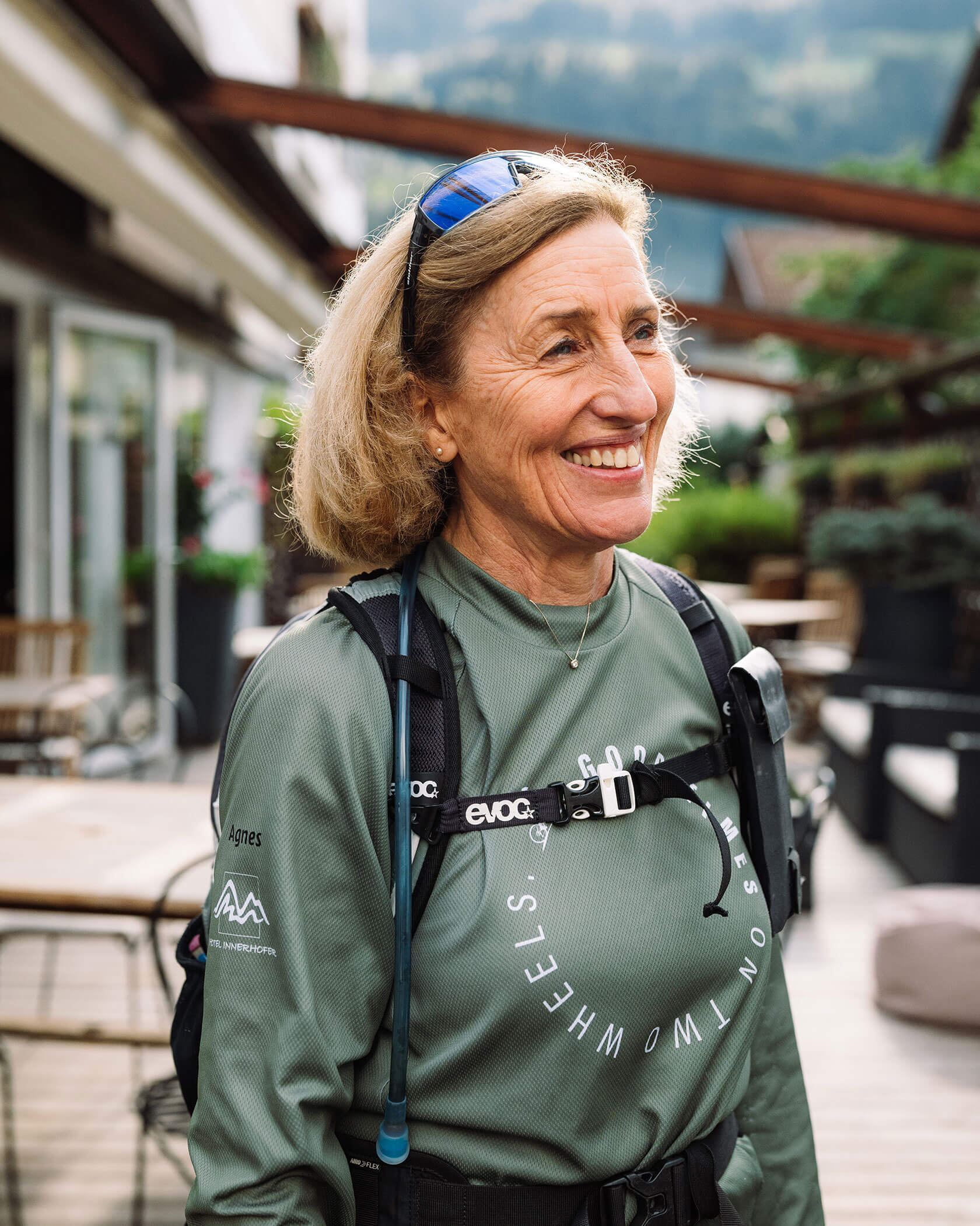 Smiling woman in outdoor gear with a backpack and sunglasses, standing on a patio with greenery behind her. - Hotel Innerhofer