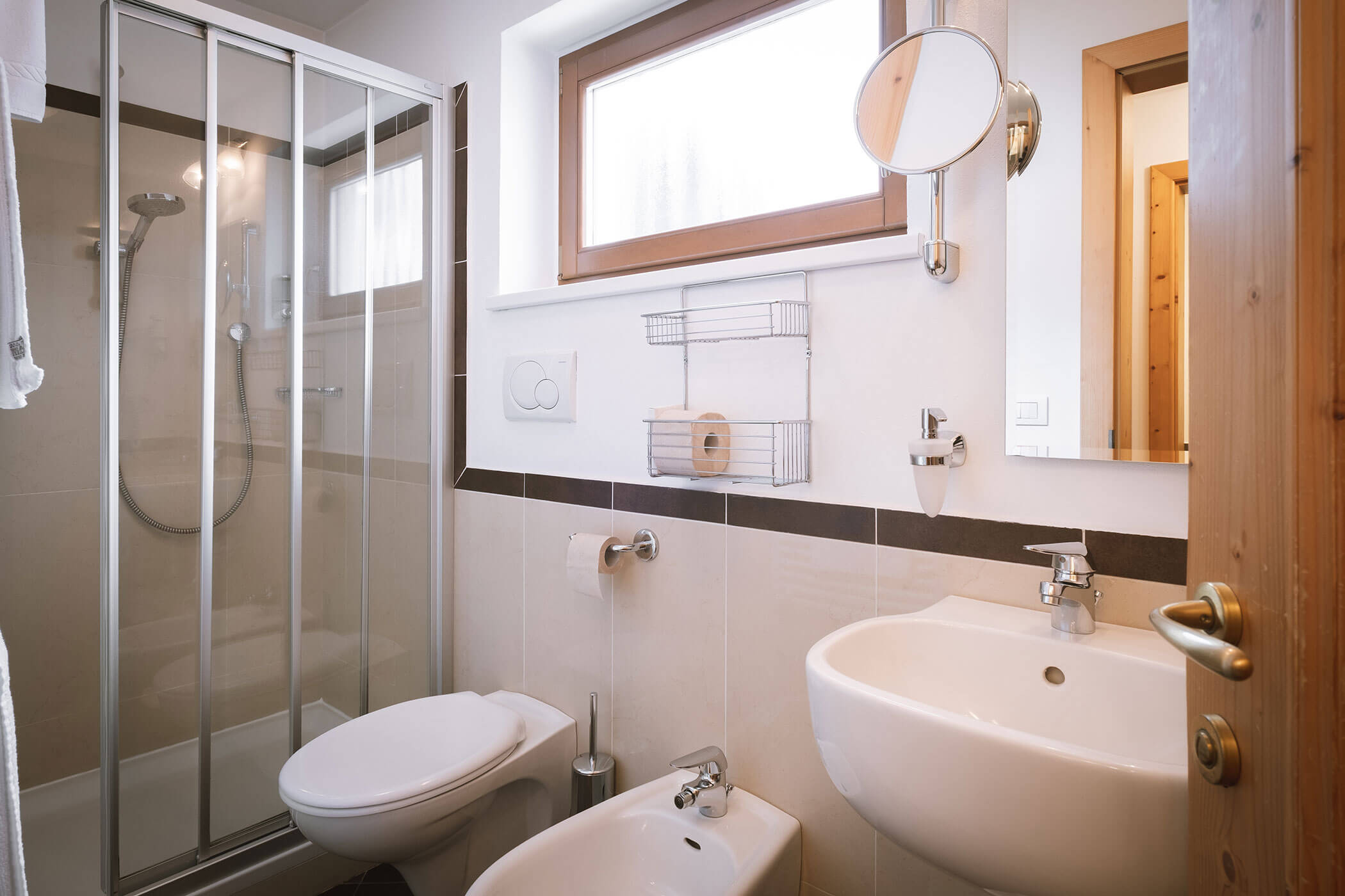 Bagno moderno con doccia in vetro, WC, bidet, lavandino bianco e finestra sovrastante. - Hotel Innerhofer