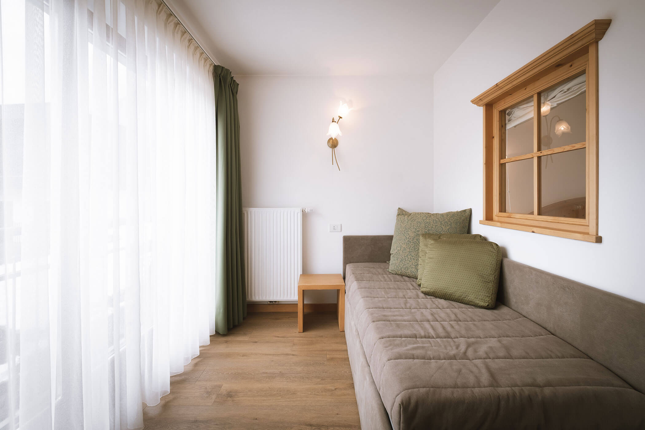 Stanza minimalista con divano marrone, cuscini verdi, pavimento in legno, ampia finestra con tende e radiatore a parete. - Hotel Innerhofer
