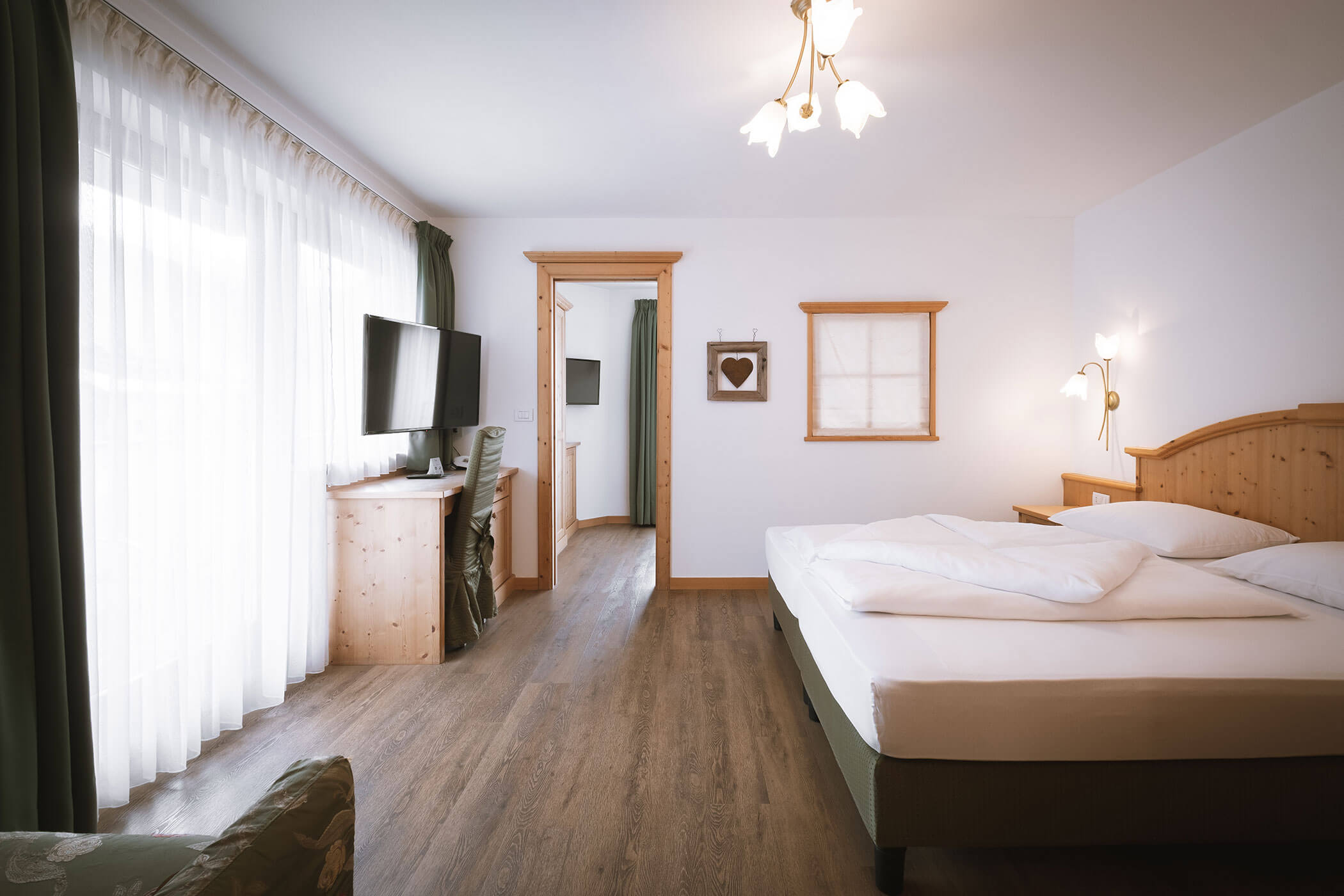 Luminosa camera d'albergo con letto matrimoniale, mobili in legno, TV, ampia finestra con tende e luci soffuse. - Hotel Innerhofer