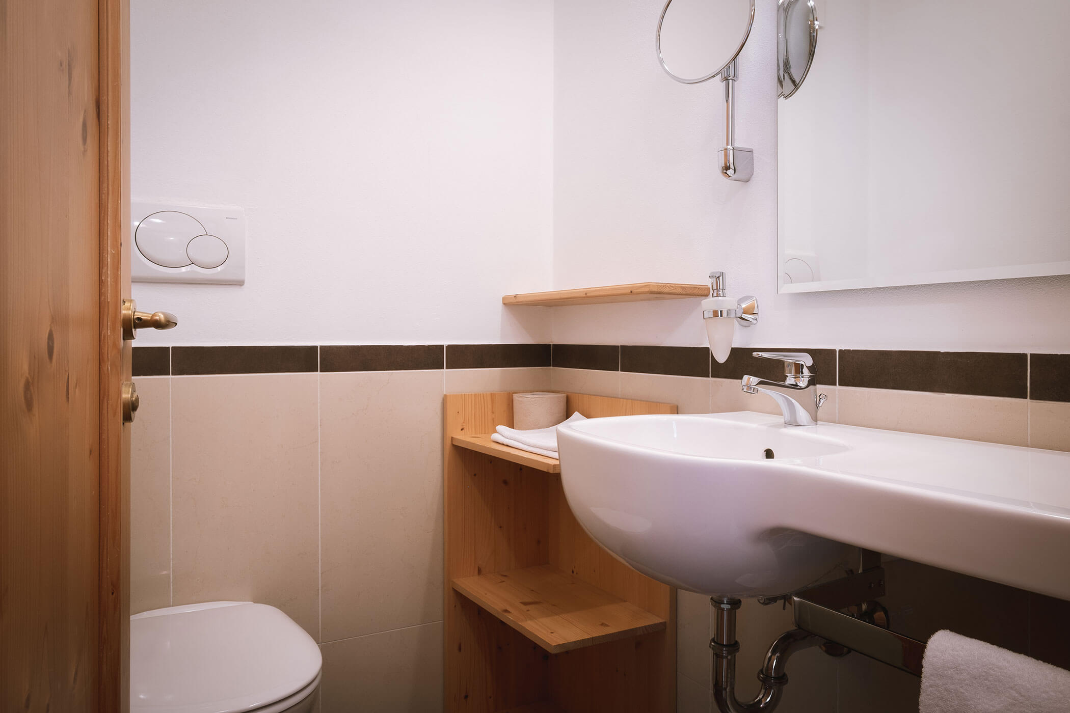 Piccolo bagno moderno con lavandino bianco, WC, scaffali in legno e specchio a parete. - Hotel Innerhofer