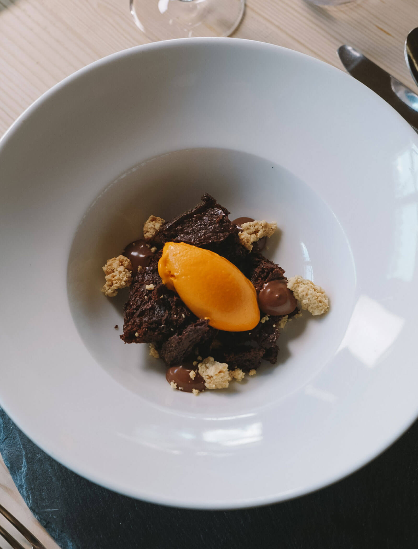 Ein Dessert mit Schokoladenkuchen, Streuseln, Schokoladensauce und einer Kugel Orangensorbet in einer weißen Schale. - Hotel Innerhofer