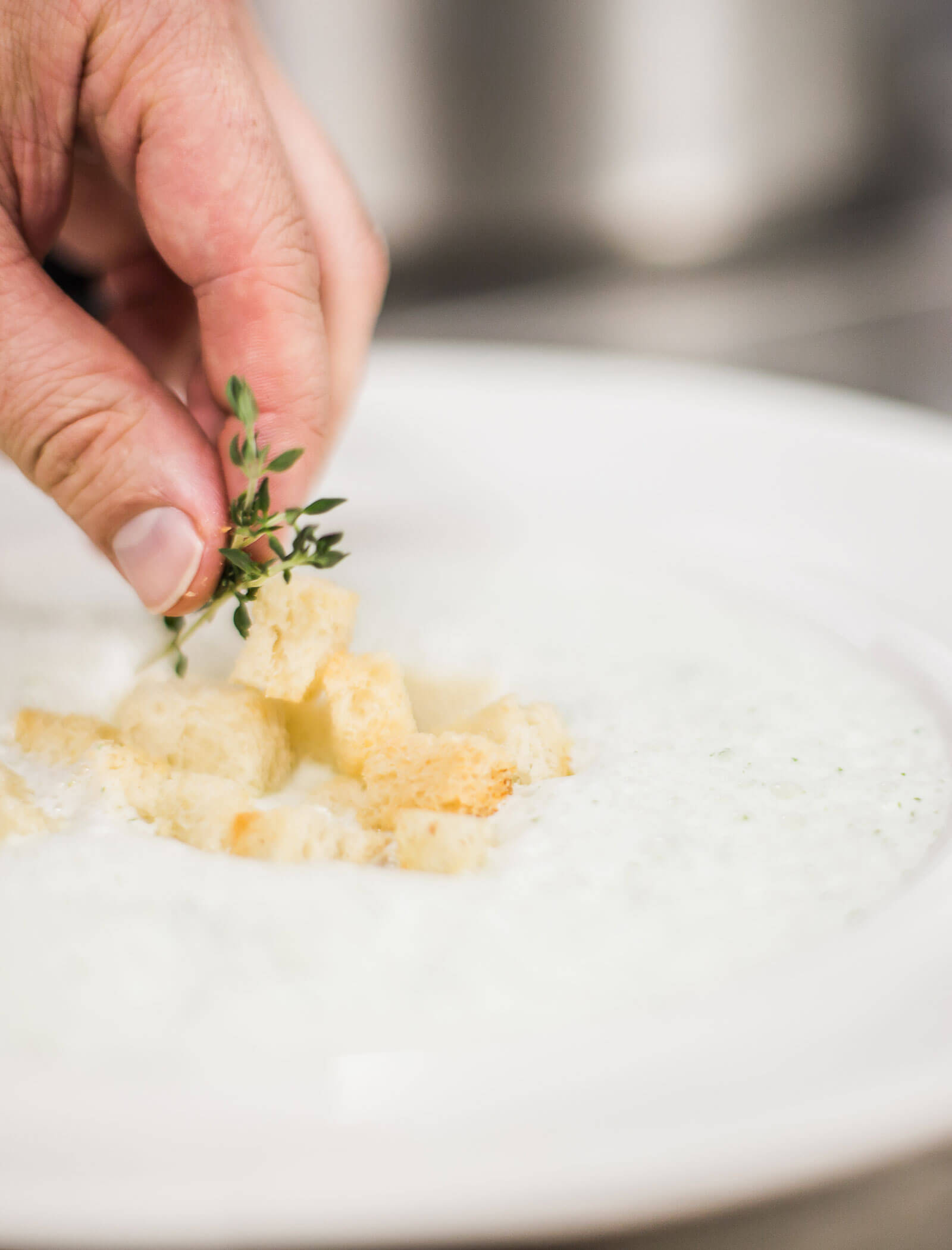 Eine Hand, die eine cremige Suppe mit Croutons und einem Zweig frischer Kräuter auf einem weißen Teller garniert. - Hotel Innerhofer