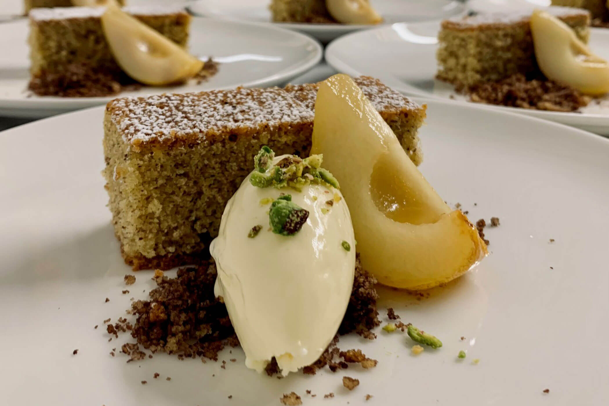 Ein Stück Kuchen mit pochierter Birne, Eis und Streuseln auf einem weißen Teller, garniert mit Pistazien. - Hotel Innerhofer