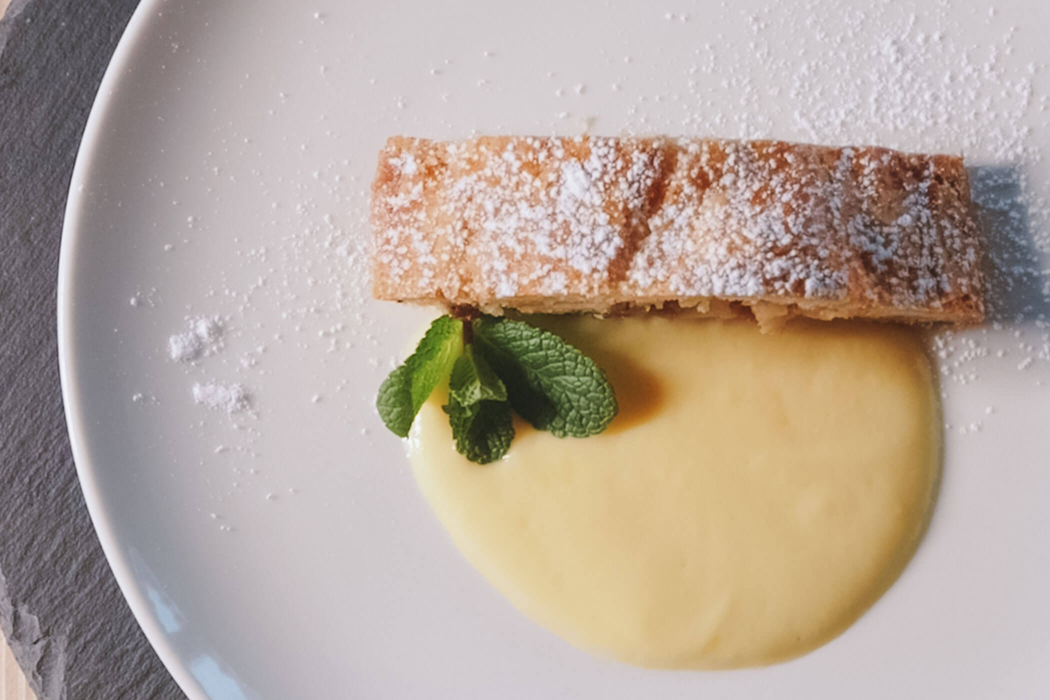 Eine mit Puderzucker bestreute Scheibe Strudel, serviert mit Vanillesoße und einem Minzzweig auf einem weißen Teller. - Hotel Innerhofer