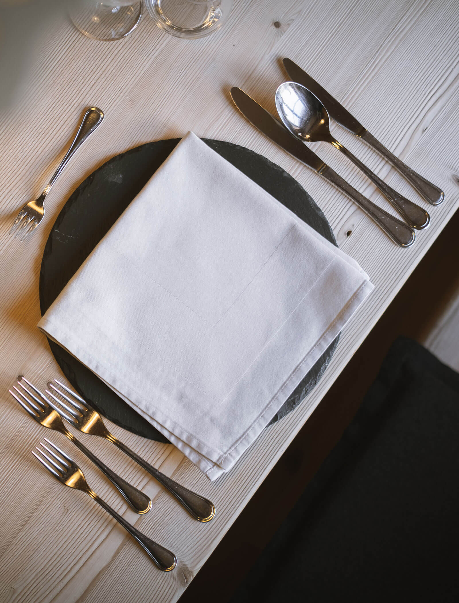 Ein gedeckter Tisch mit Silberbesteck, einem runden dunklen Teller und einer weißen Serviette auf einem hellen Holztisch. - Hotel Innerhofer