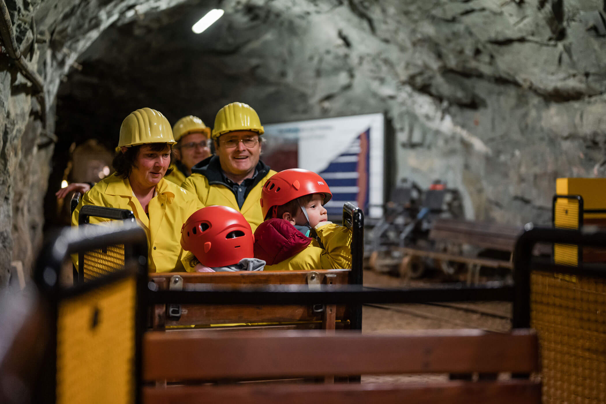Adults and children in yellow helmets and jackets sit in a mining cart inside an underground tunnel. - Hotel Innerhofer
