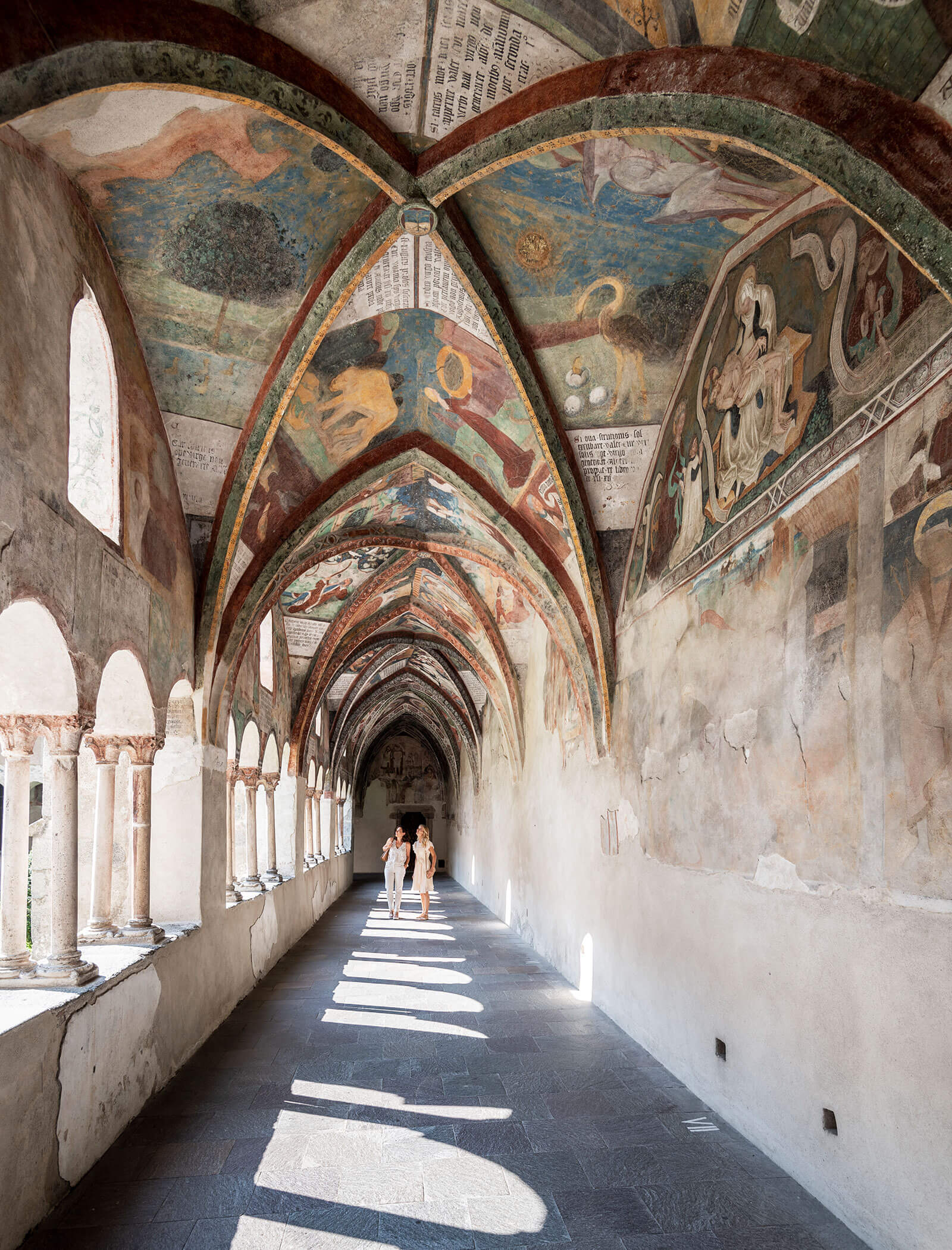 People walking through a sunlit cloister with arched ceilings and colorful frescoes on the walls and ceiling. - Hotel Innerhofer