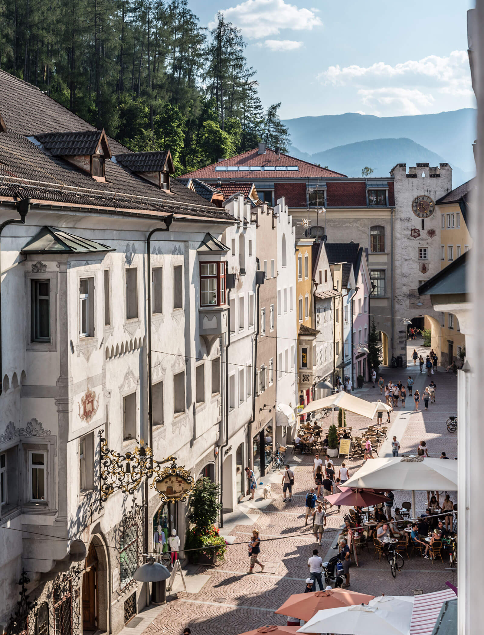 Sunlit European street with old buildings, outdoor cafes, people walking, and mountains in the background. - Hotel Innerhofer