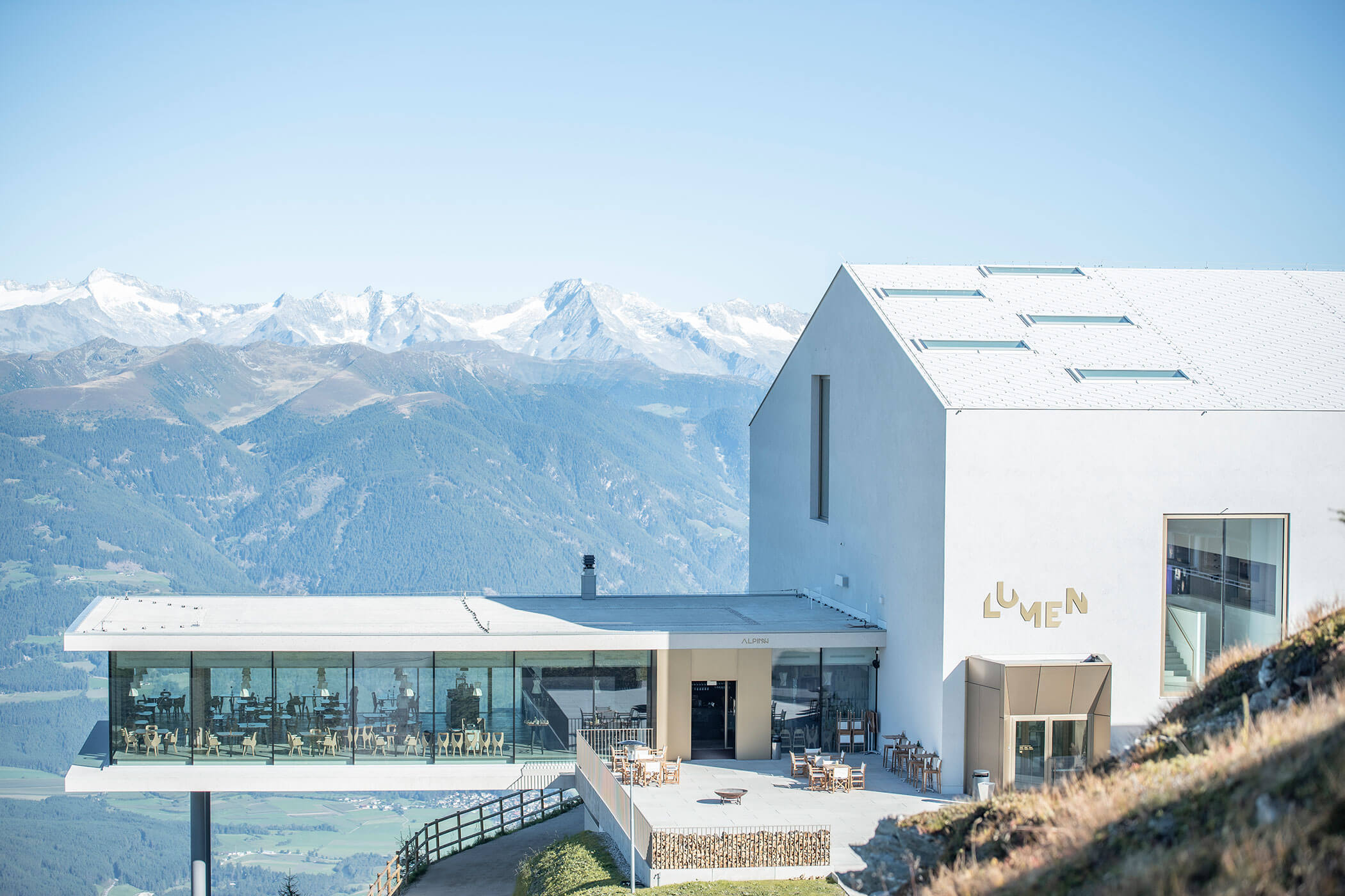 Modern mountainside restaurant with large glass windows, overlooking scenic snowy peaks and valleys under a clear sky. - Hotel Innerhofer