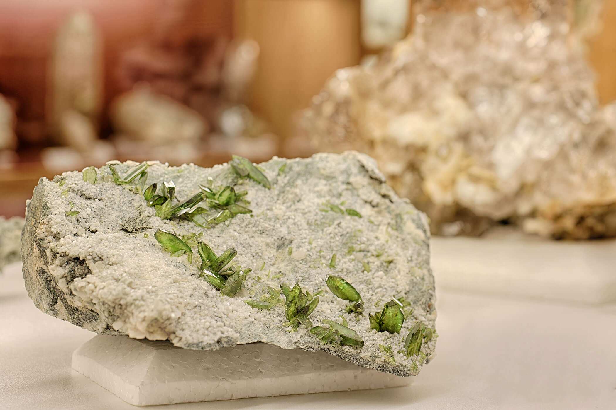 A rough mineral rock with green crystal formations displayed on a white platform indoors. - Hotel Innerhofer