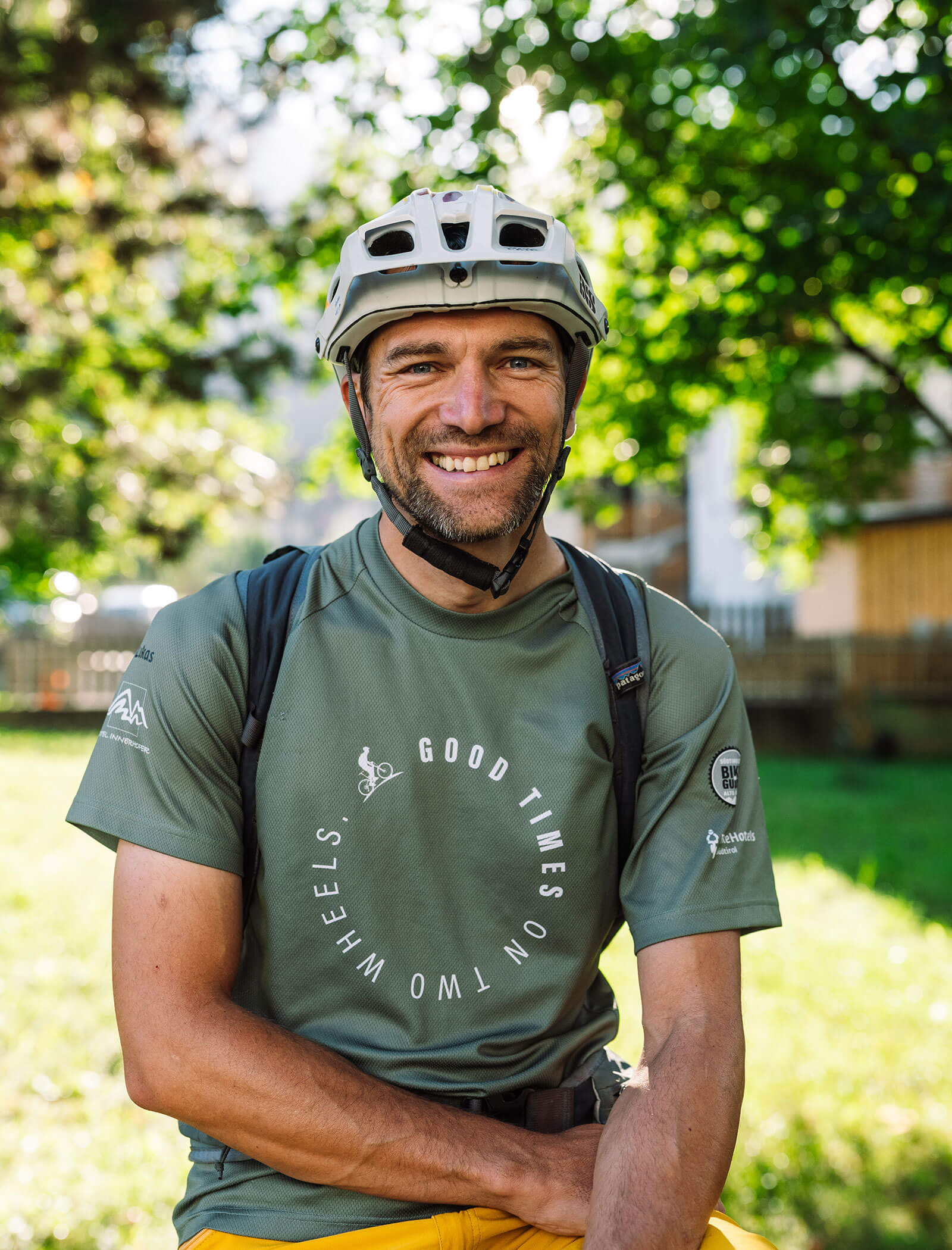Un uomo sorridente con casco da bici e camicia verde siede all'aperto in una giornata di sole con alberi sullo sfondo. - Hotel Innerhofer