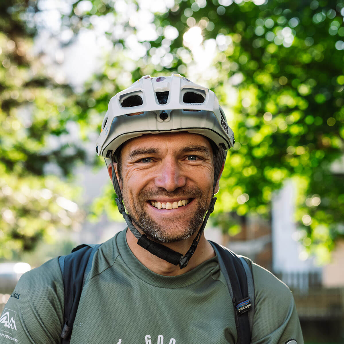 Un uomo sorridente con casco da bici e zaino in spalla si trova all'aperto con alberi e luce del sole sullo sfondo. - Hotel Innerhofer
