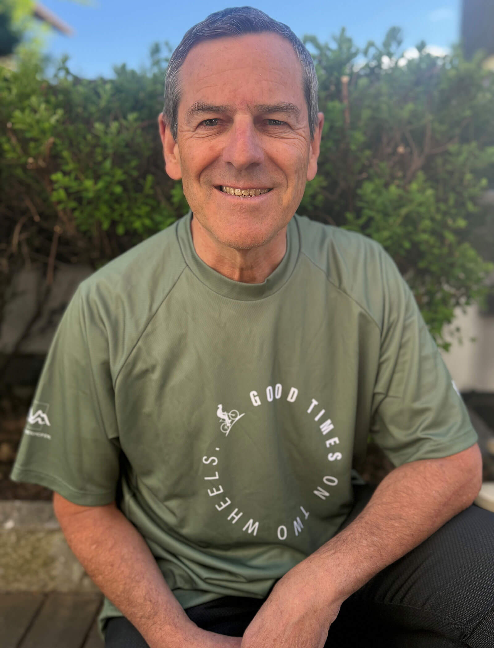 Un uomo anziano e sorridente con una maglietta verde di Good Times On Two Wheels siede all'aperto davanti a cespugli verdi. - Hotel Innerhofer
