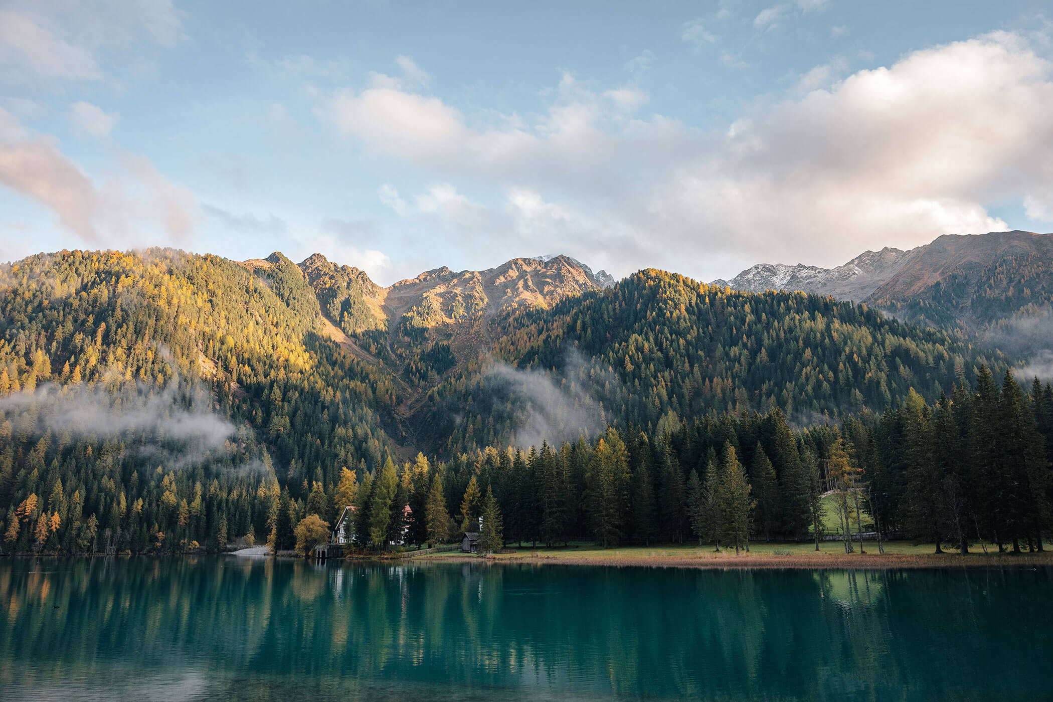 Clear lake with reflections, dense forest, and sunlit mountains under a partly cloudy sky. - Hotel Innerhofer