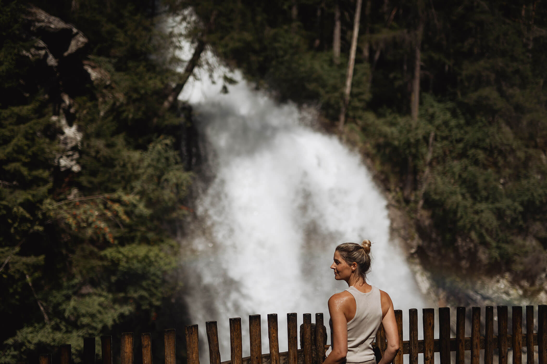 Woman in a white tank top stands by a wooden fence, looking at a waterfall surrounded by trees. - Hotel Innerhofer